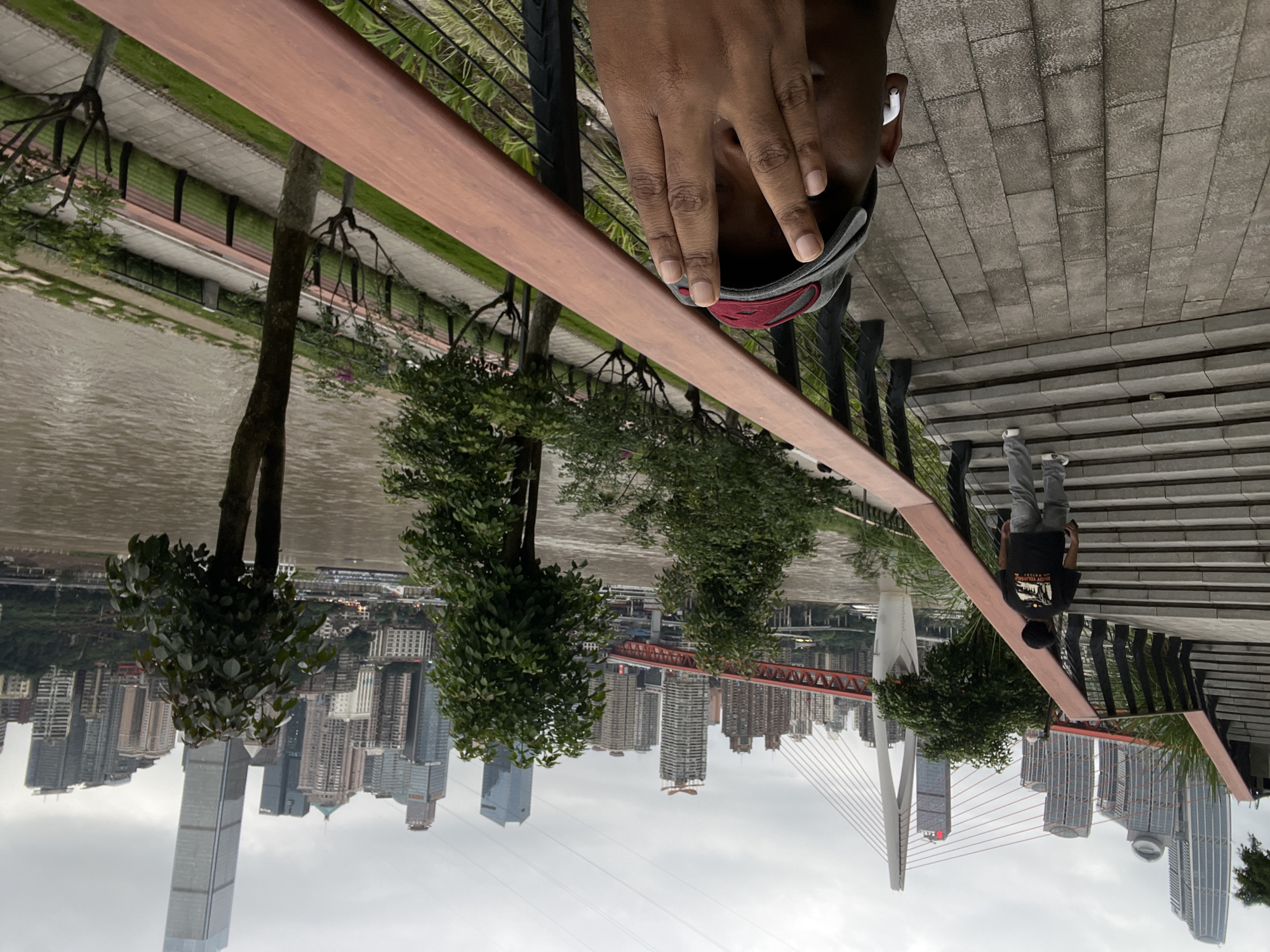 A person with dark skin is leaning against a wooden railing, his face partially obscured by his hand, with an earbud visible in his ear. Another person is walking down a set of stairs to the right. Below the railing, there is a river or body of water. On the opposite side of the water, there is a cityscape with numerous tall buildings and a distinctive bridge with a white tower. The photograph was taken in 江北城街道, China.