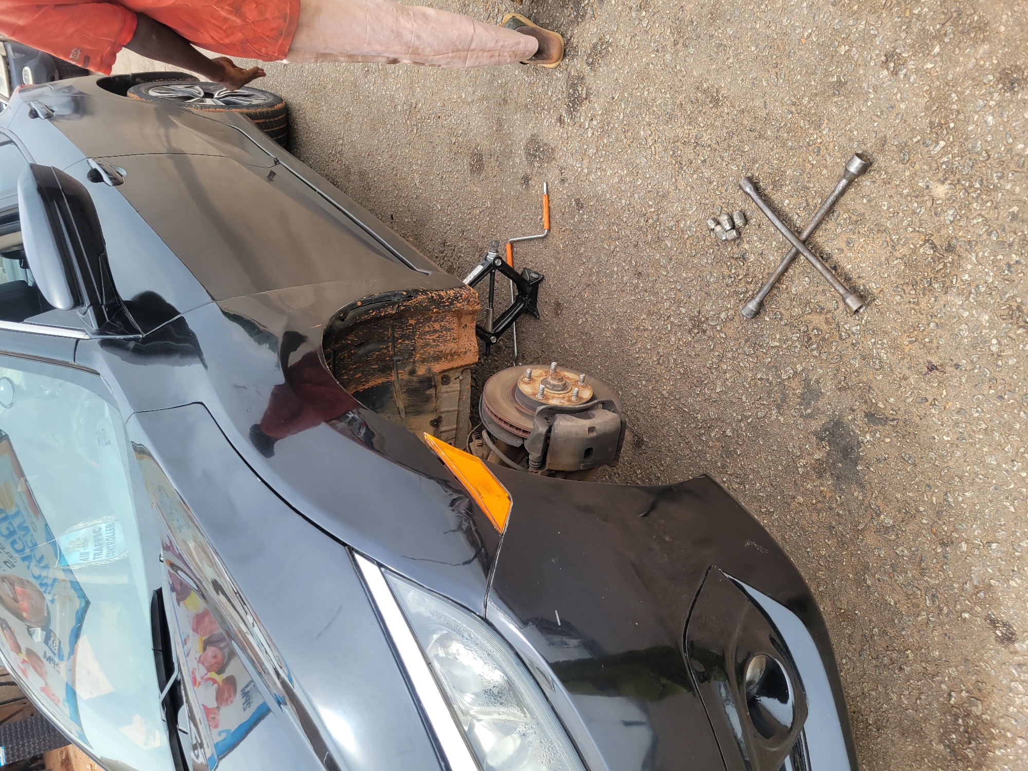The image shows a black car with its front passenger-side wheel removed, revealing the brake rotor, caliper, and suspension components. The car is lifted by a black scissor jack. A lug wrench and several lug nuts are on the ground. Part of a tire is visible next to the car. A person wearing an orange outfit and brown footwear stands nearby. The location is outdoors on a paved surface. The city and state are unknown.