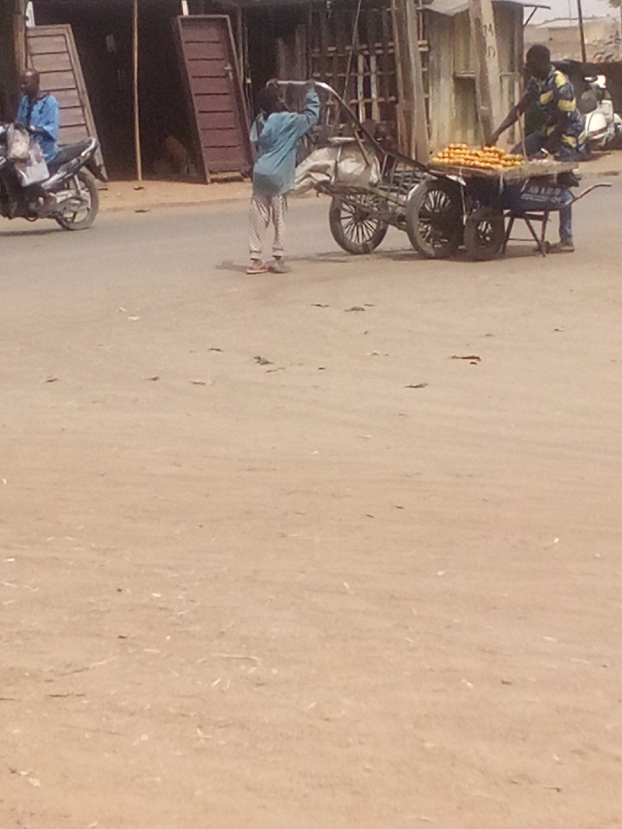 The image depicts an outdoor scene on a dusty, light brown ground surface in Tudun Murtala, Nigeria. Three individuals are present. On the far left, a man in a light blue long-sleeved shirt and blue trousers sits astride a dark-colored motorcycle, which has a white bag on its seat. In the center, a person wearing a light blue long-sleeved top and patterned light-colored trousers stands facing away from the viewer, holding the elevated handle of a two-wheeled handcart equipped with large wooden spoked wheels and carrying a light-colored sack. To the right, a man in a patterned yellow, blue, and white shirt stands behind a four-wheeled cart displaying numerous rows of orange fruits. The background features several simple structures, some with dark wooden doors or shutters and visible metal grates.