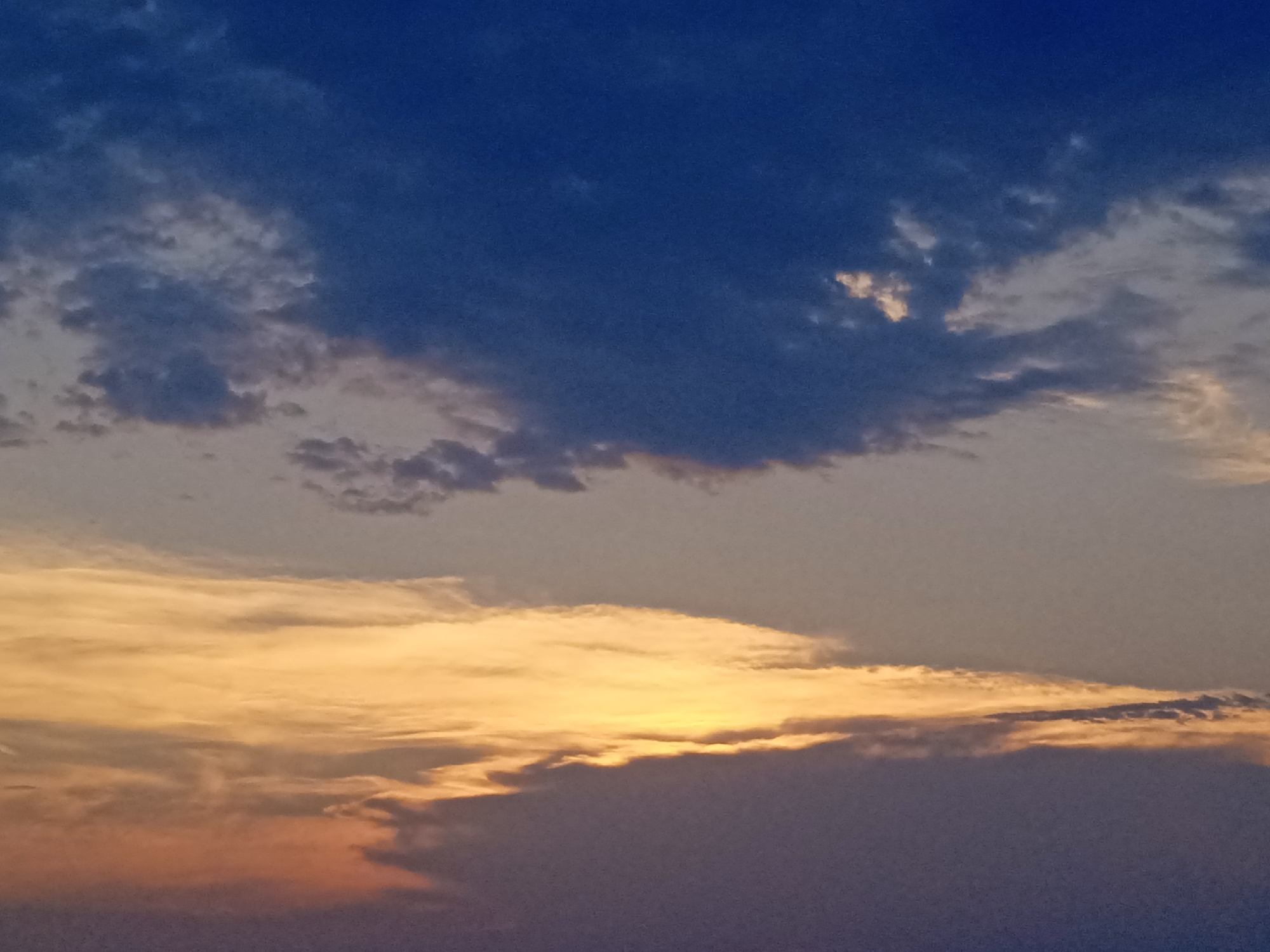 The image is a low-angle, medium shot of a twilight sky filled with clouds. The sky transitions from a deep, dark blue at the top to lighter shades of gray and yellow towards the bottom. Wispy, dark gray clouds are scattered across the blue expanse, with some lighter, almost white clouds interspersed. The lower half of the sky is dominated by a wide band of bright, golden-yellow clouds, tinged with orange and pink at the edges. The very bottom of the image is a dark, indistinct mass, possibly representing distant land or clouds. The lighting suggests either sunrise or sunset. The image does not depict any specific city or location.
