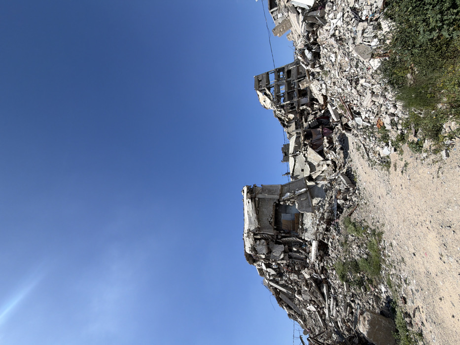 The image depicts a scene of extensive urban destruction in Jabalia, Palestinian Territory, under a clear blue sky with a sun flare visible in the upper left. The foreground features a light-colored dirt path or track, veering slightly to the right, with sparse green vegetation, including weeds and small bushes, growing along its edges and within the surrounding debris.
The middle ground and background are dominated by massive piles of rubble, remnants of what appear to be multi-story concrete buildings. On the left, a large mound of pulverized concrete, twisted rebar, and building fragments rises, with a partially intact, possibly internal, room structure visible within the debris. To the right, another significant mound of similar debris features the skeletal remains of a building, showing multiple exposed floor plates and support columns, indicative of severe structural damage and partial collapse. Exposed internal elements, such as damaged walls and fragments of personal belongings (e.g., fabric), are discernable within the shattered structures.
The entire landscape is characterized by widespread destruction, with no individuals visible within the frame. The scene is illuminated by strong sunlight, casting minimal shadows due to the overhead sun.
