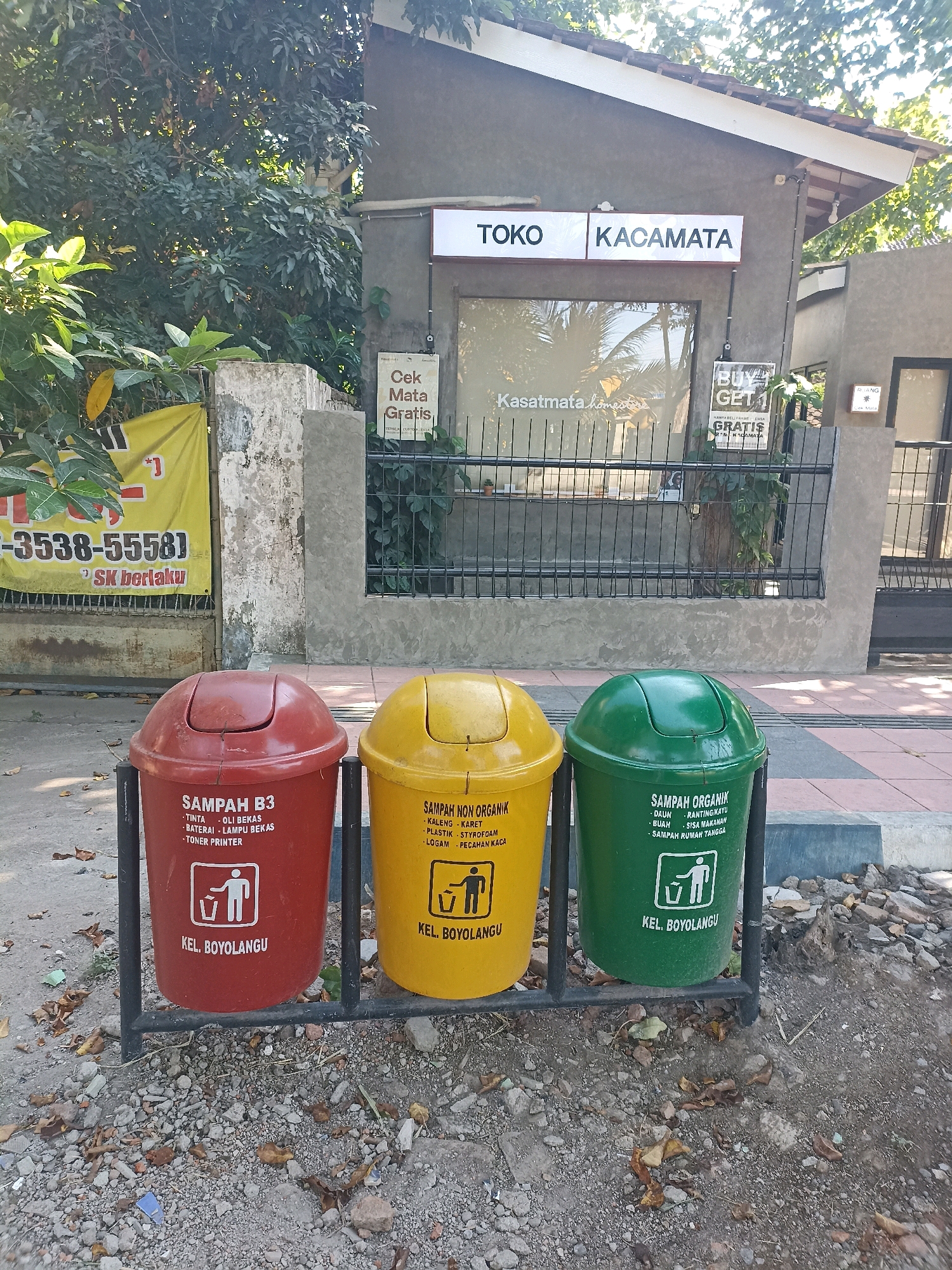 The image shows a row of three colored trash cans in front of a building with a sign reading "TOKO KACAMATA" in Mojopanggung, Indonesia. The trash cans are red, yellow, and green, and each has labels indicating what type of waste they are for. The red can is for "SAMPAH B3" (Hazardous and Toxic Waste) and lists items like ink, oil, batteries, lamps, and printer toner. The yellow can is for "SAMPAH NON ORGANIK" (Non-Organic Waste) and includes items like plastic, styrofoam, and broken glass. The green can is for "SAMPAH ORGANIK" (Organic Waste) and lists items such as leaves, wood, fruit, food scraps, and household waste. Below these categories, each bin has a pictogram of a person throwing trash and the text "KEL. BOYOLANGU". The building behind the trash cans has a glass window with "KASATMATA HOMES" written on it. There is also a sign with "BUY 1 GET 1 GRATIS" suggesting a promotion. The environment appears to be outdoors, with trees and foliage visible in the background. The ground is a mix of dirt, gravel, and fallen leaves.