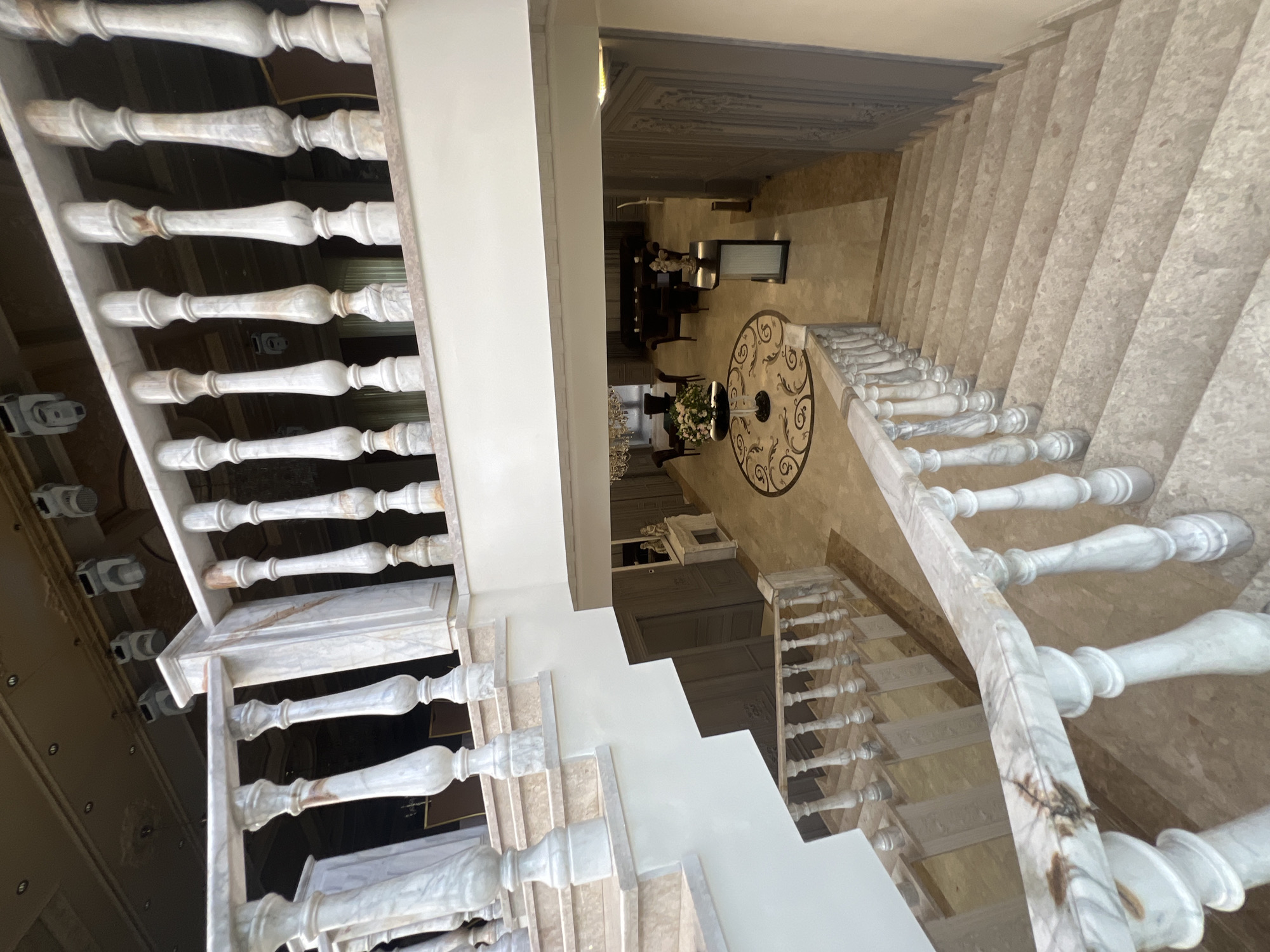 The image displays a grand, opulent interior space, likely a hall or foyer, viewed from an upper floor looking down onto a sweeping staircase and the floor below. The photograph is oriented vertically, showing the staircase descending from the left towards the right.
The central subject is a multi-turn staircase made of light-colored, possibly beige or cream-toned marble or polished stone. The steps are wide and substantial. The balustrade is crafted from white and grey veined marble, featuring numerous elegantly turned balusters supporting a polished marble handrail. A landing is visible in the middle section of the stairs.
The floor of the hall below is paved with a light, reflective stone, possibly marble or travertine. A striking feature is a large circular inlaid medallion in the center of the floor, showcasing an intricate, dark, swirling floral or scroll pattern against a lighter base.
Around the medallion, classic-style dark wooden chairs are arranged around tables. One table in the foreground holds a large, elaborate floral arrangement with white and pink blooms. Further back, another console table or buffet is visible with decorative objects. A large, ornate chandelier hangs partially visible above the central area.
The ceiling of the hall features elaborate decorative moldings or panels, painted in a light, possibly grey, hue, contributing to the luxurious aesthetic. Dark wooden archways or doorways can be seen in the background, leading to other rooms. The lighting suggests a well-lit interior, either from natural light entering from unseen windows or sophisticated artificial lighting. No people are present, and no specific activity is taking place. There is no visible text. The overall impression is one of classical elegance and grandeur within a spacious setting in Yerevan, Armenia.
