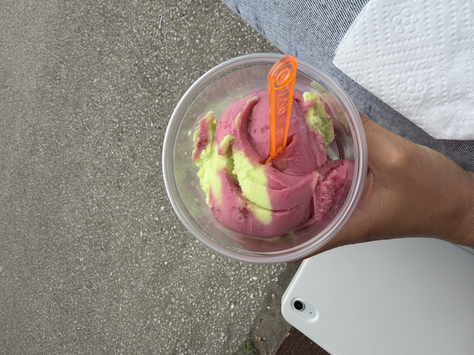 An outdoor, overhead shot captures a person's hand holding a clear plastic cup filled with two-toned ice cream, a swirl of pink and light green. An orange plastic spoon is nestled in the ice cream. To the left of the cup, a white tablet lies on a wooden surface. In the background, a grey, speckled asphalt surface suggests an outdoor setting, possibly a public space in Pazardzhik, Bulgaria. The person's legs, clad in blue jeans, are visible on the right side of the frame, along with a white paper napkin with a textured pattern.