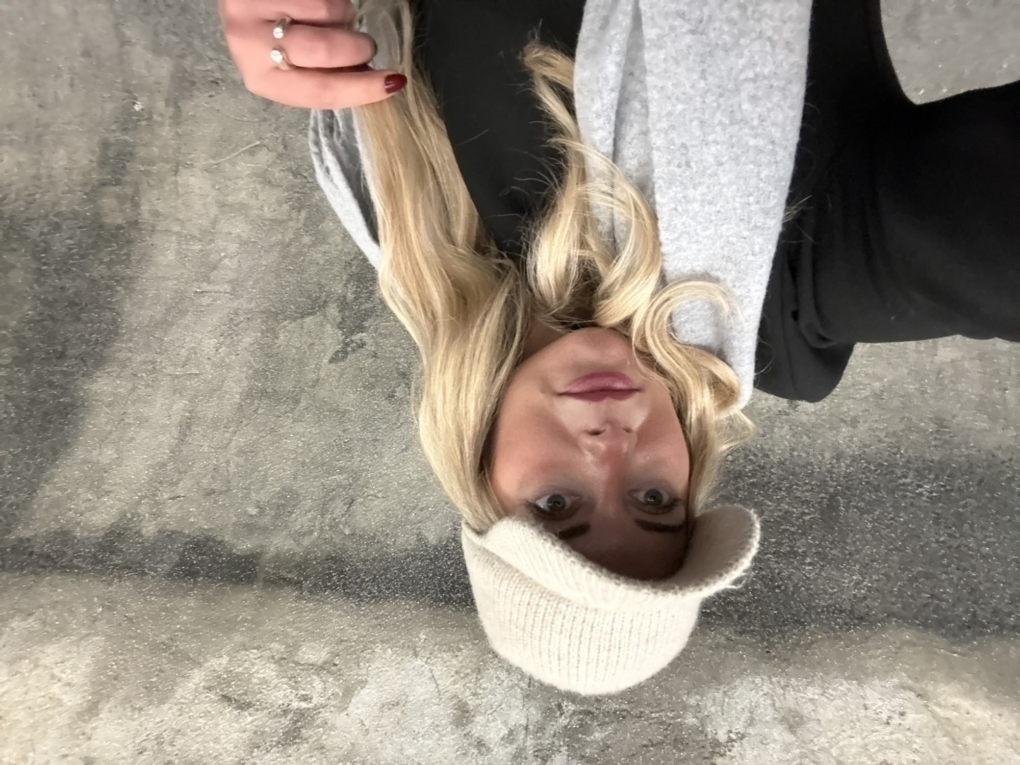 A woman wearing a white knit hat, grey scarf, and black top is lying on a grey concrete surface. She has blond hair and is wearing rings on her left hand. The background appears to be a city sidewalk or similar paved area. There is no indication of location in Paris.