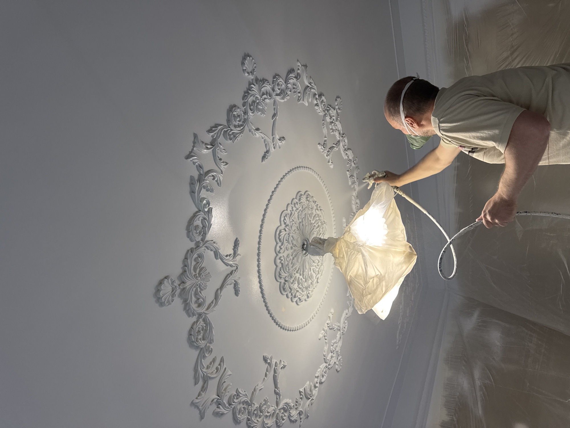 A man wearing a respirator is spray-painting a ceiling with ornate plasterwork in a room in the city of Chaikovsky, Russia. The ceiling has a circular design around a light fixture that is covered with a protective cloth. The walls of the room are covered with plastic sheeting. The man is holding a spray gun with a hose.