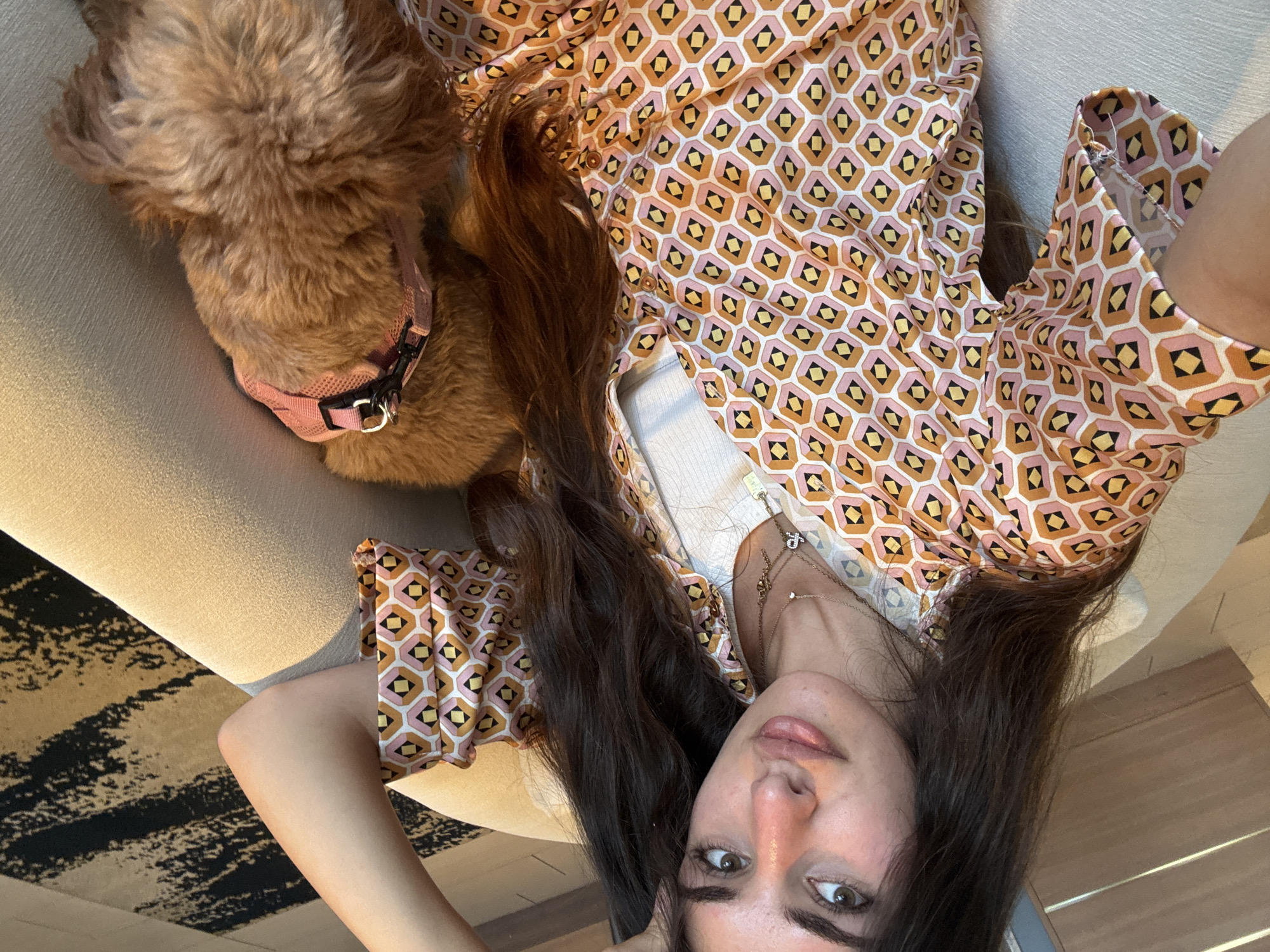 In a comfortable indoor setting, a woman with long, dark hair is seated on a beige couch, her right arm resting on her head. She wears a collared, short-sleeved shirt with a geometric pattern of pink, orange, and yellow hexagons with black and white diamond accents. Underneath, a white tank top is visible. She has a subtle smile and is looking towards the camera. Next to her, a small, fluffy, reddish-brown dog is lying down, wearing a pink harness. The dog's head is lowered, obscuring its face. The background includes a wooden wall with horizontal slats and a patterned rug with black and beige stripes. The scene appears to be in a residential living room in Boca Raton, Florida, USA.