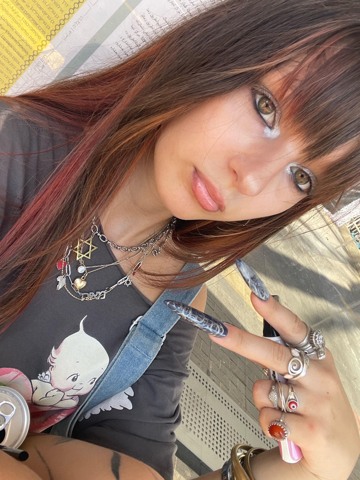 A young woman with long, auburn-streaked brown hair and bangs poses for a selfie outdoors. She wears a dark gray t-shirt with a graphic of a cartoon baby, a denim strap across her chest, and several layered silver necklaces, including a Star of David pendant. She has dramatic white eyeliner and prominent rings on her fingers, as well as long, dark, patterned artificial nails. Her left hand is raised, showing off her nails and rings. A silver soda can is visible to the left of her torso. The background appears to be an outdoor urban environment with a tiled walkway and a yellow and white patterned structure that might be part of a public transportation ticket or map. The lighting suggests it is daytime.