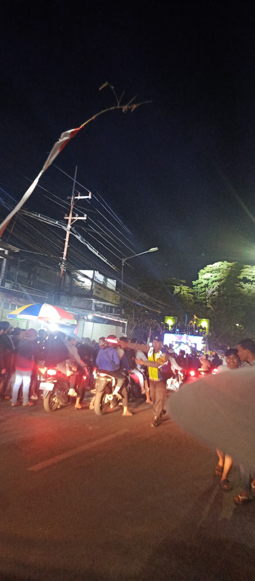 A nighttime street scene in Sekarpuro, Indonesia, shows a crowd of people and several motorcycles. A traffic officer in a yellow and black vest stands on the right, appearing to direct traffic or manage the crowd. The street is illuminated by streetlights, vehicle lights, and possibly lights from a nearby event or stage, which is visible in the background with large speakers and colorful lights. Some individuals are on motorcycles, while others are standing or walking. A striped umbrella is visible on the left side of the image. Overhead power lines and utility poles are present, typical of an urban Indonesian environment. The overall atmosphere suggests a lively public gathering or event.
