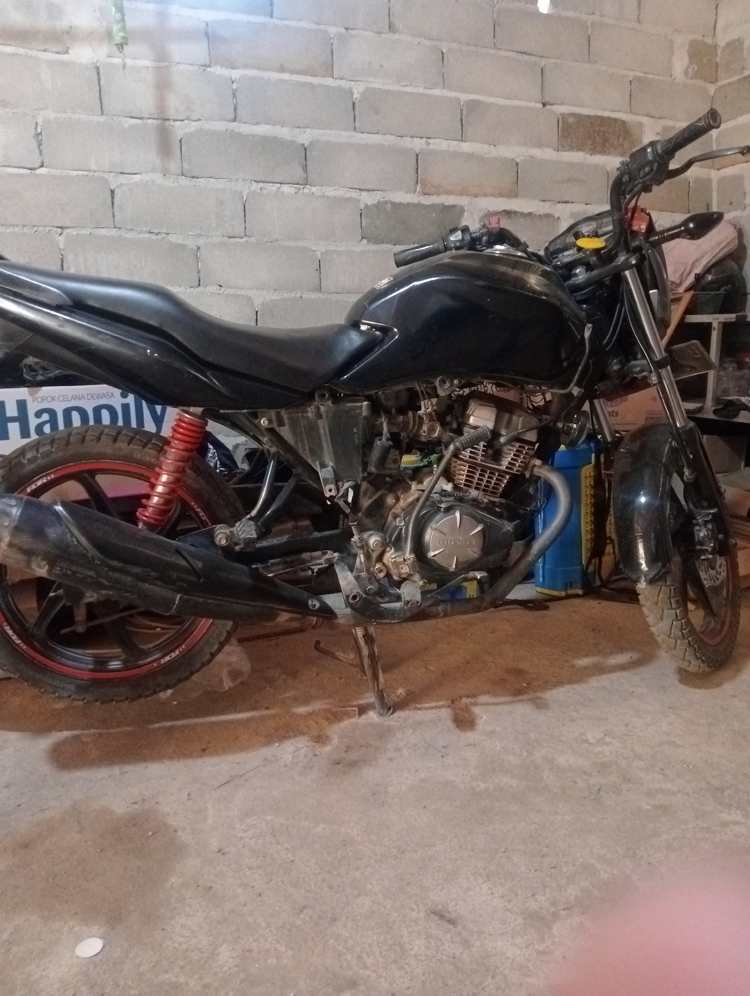 An old black motorcycle is parked in a concrete garage with a cinder block wall in the background. The motorcycle is facing right, and its side is visible. The motorcycle has a worn-out appearance, with some parts showing rust and dirt. The red shock absorber and red rim on the rear wheel are noticeable. A blue container and other items are behind the motorcycle. The floor is concrete and appears to be stained. The scene is likely a residential garage in Kota Negara Timur, Indonesia.