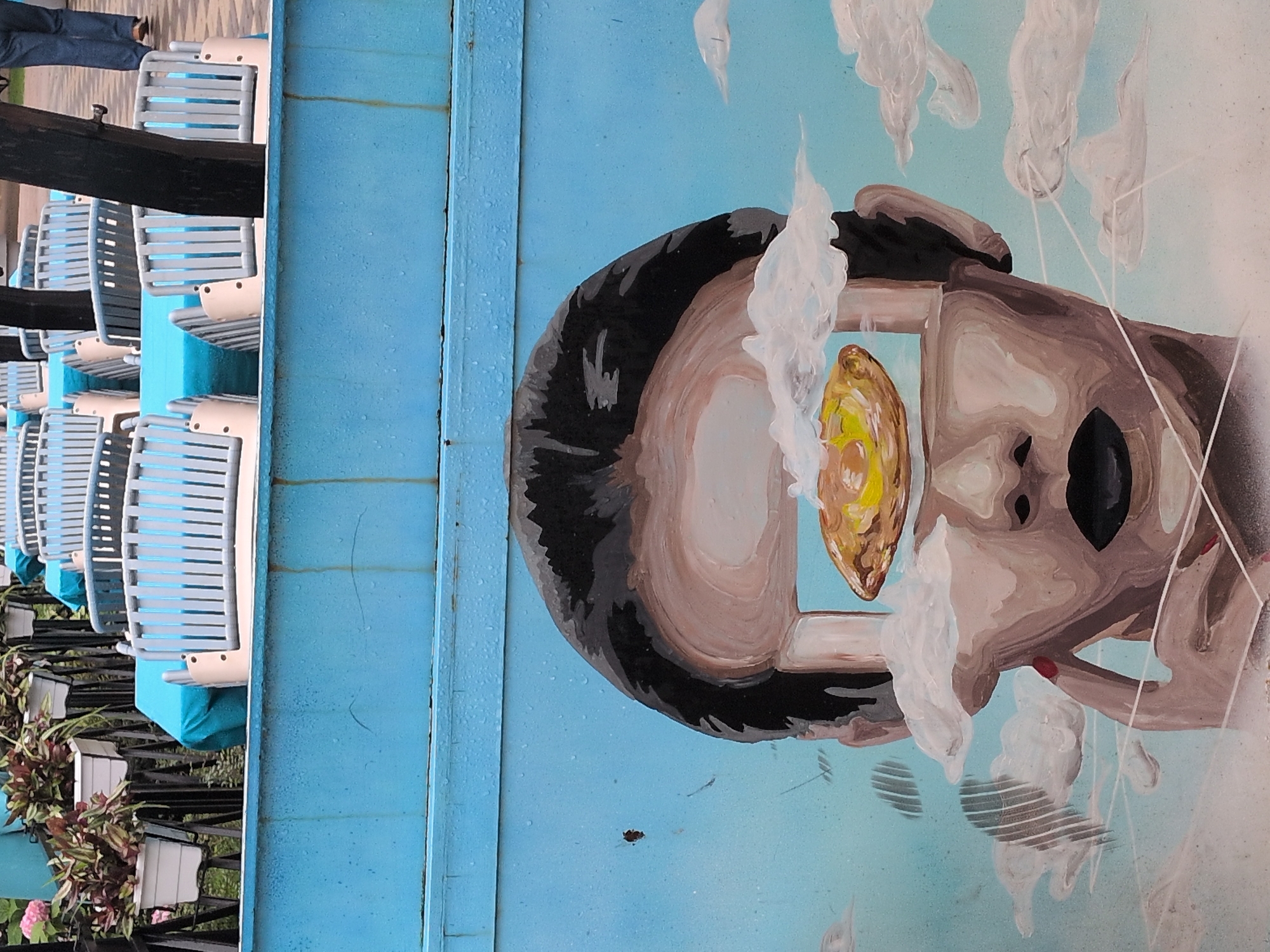 The image displays a street art mural of a face with an abstract element in its forehead, set against a blue sky with clouds. The mural is located on a light blue painted surface, possibly a wall or barrier. In the background, rows of empty light blue chairs are visible, some covered with blue tarps. Potted plants with red and green foliage are also present. The overall setting appears to be an outdoor public space. The mural is detailed and vibrant, featuring a woman's face with black lips, rendered in a surreal style. The surrounding environment suggests a recreational area or an outdoor seating space in Sochi, Russia.