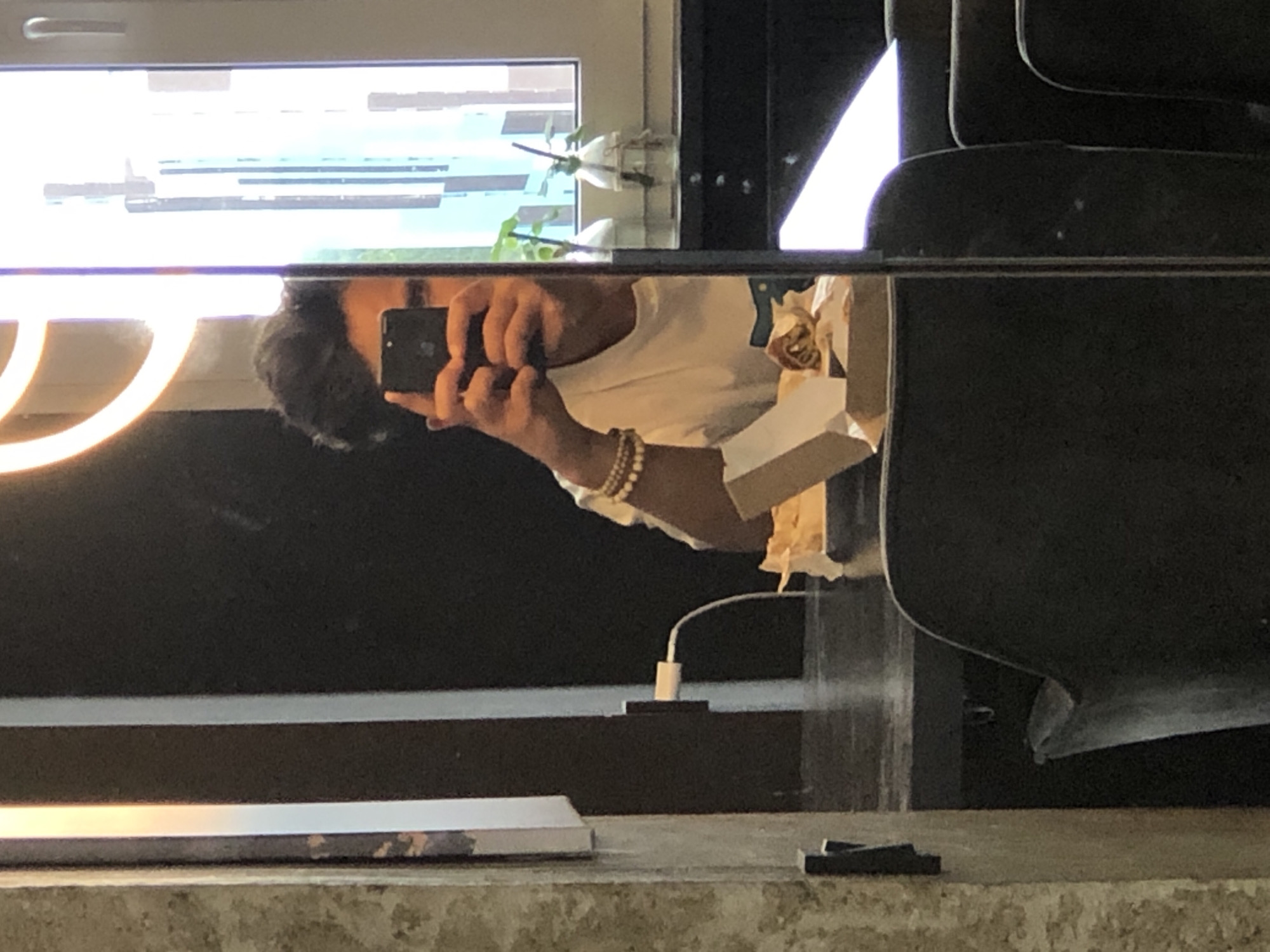 In Saint Petersburg, Russia, a man is reflected in a mirror, taking a selfie with his smartphone. He is wearing a white t-shirt and several beaded bracelets on his left wrist. In front of him, on a dark countertop, is an open takeout box containing food. Behind him, a power adapter is plugged into a white wall socket, with its cable trailing away. The man's dark, messy hair is visible. The background of the reflection shows a window with blinds and two small potted plants in glass vases. The overall setting appears to be an indoor cafe or restaurant, with dark seating visible in the foreground.