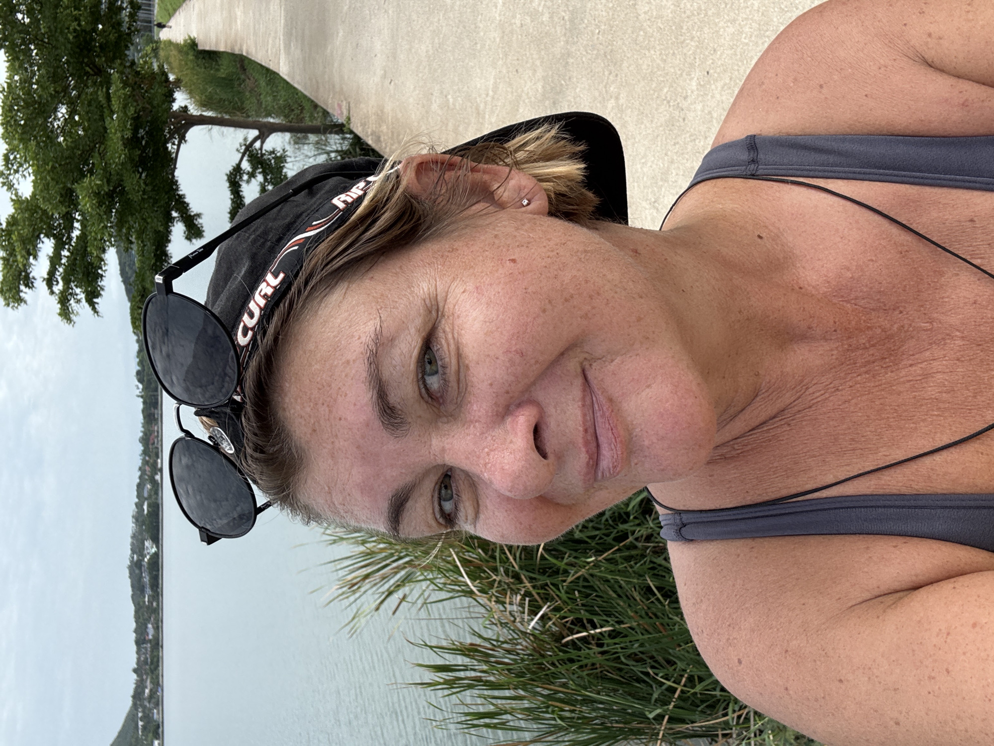 A woman is standing outside near a body of water in Baan Chaweng, Thailand. She is wearing a gray tank top and a black baseball cap with sunglasses perched on top. Green vegetation is visible near the water's edge, and a paved walkway and trees are in the background. The sky is overcast.