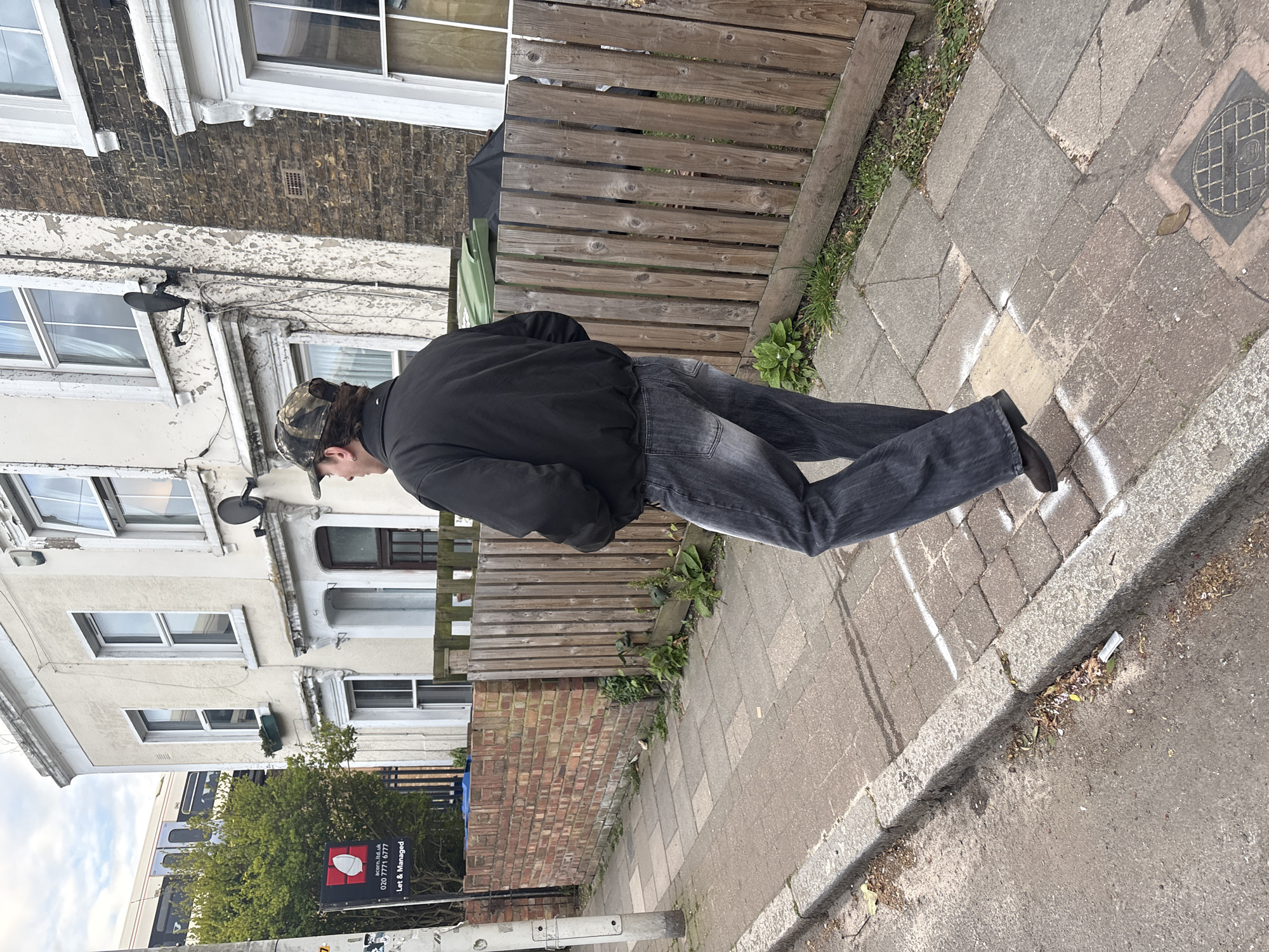 The image depicts a residential street scene in London, United Kingdom, under an overcast sky. A male subject, seen from behind, walks rightward on a paved sidewalk. He wears a black bomber jacket, dark grey jeans, and a camouflage baseball cap worn backward; a silver stud is visible in his left ear. Two distinct white spray-painted rectangular marks are visible on the concrete slab sidewalk. A grey curb separates the sidewalk from a dark asphalt street. To the right, a weathered wooden picket fence and a low brick wall separate the public thoroughfare from properties, with weeds growing along their bases. Behind the fence, a green waste bin and a black tarp-covered item are visible. Beyond, terraced houses line the street, featuring both light-colored stucco and dark brick facades with white window frames; one has a small black satellite dish. A "Let & Managed" sign for "acorn.ltd.uk" (020 7771 8777) is affixed to a pole on the left. In the background, a white and blue commuter train passes on an elevated track, partially obscured by trees.