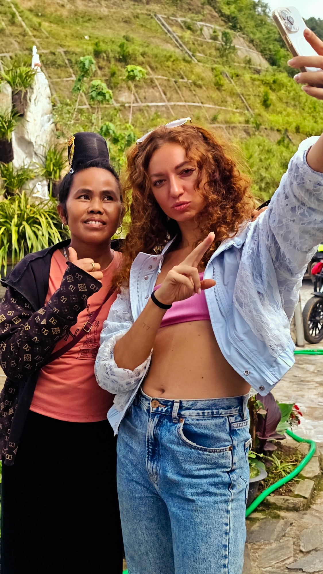 This image, captured in Tu Le, Vietnam, depicts two women posing for a selfie outdoors during daylight. On the left, a woman of apparent Southeast Asian descent, likely in her 30s or 40s, smiles directly at the camera and gives a thumbs-up gesture with her right hand. She has dark hair styled in a tall, elaborate bun with a gold-colored ornament. Her attire includes a coral-pink t-shirt, a black jacket featuring a repeating light-colored 'DG' monogram pattern on the sleeves, and black lower garments. A purple strap is visible across her chest. On the right, a woman of apparent Caucasian or Mediterranean descent, likely in her 20s or 30s, holds a light-colored smartphone with her raised left hand, taking the selfie. She has long, curly, reddish-brown hair with sunglasses perched on her head. Her expression features pursed lips, and she gestures forward with her raised right index finger. She wears a pink bandeau top, a light blue lace-textured jacket, and light blue denim jeans, exposing her midriff. A '77' tattoo is visible on her right forearm, and a black elastic band is on her right wrist. The background consists of a verdant, terraced hillside under bright sunlight, interspersed with lush green foliage. A white stone structure with surrounding plants is visible on the far left. The foreground shows a stone or paved pathway, a green hose, and parts of a motorbike on the right.