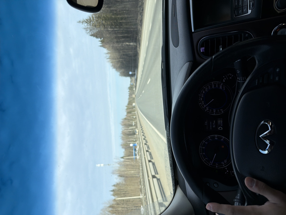 The image displays a first-person perspective from the driver's seat of an Infiniti vehicle, traversing a multi-lane highway in the vicinity of Ivanovka, Russia. A driver's left hand is positioned on the black leather steering wheel at the 9 o'clock position, featuring a silver Infiniti emblem. The instrument cluster indicates the vehicle is in 'D' (Drive), with the speedometer showing approximately 100-110 km/h and the tachometer at roughly 2000-3000 RPM. The digital odometer reads 199,026 km, with a trip meter displaying 52.1 km, and an external temperature of 5°C.
Ahead, the asphalt highway extends linearly under a partly cloudy, light blue sky. Both edges of the highway are bordered by metal guardrails and then by dense formations of leafless deciduous trees, suggesting a late winter or early spring environment. On the left side of the highway, a tall white cellular tower is prominent, alongside a small blue-roofed building and several parked vehicles, indicative of a roadside stop or service area. Multiple other vehicles are visible in the distance on the highway. The upper portion of the windshield features a dark blue tint, and a segment of the vehicle's dashboard and the rearview mirror are visible on the right.