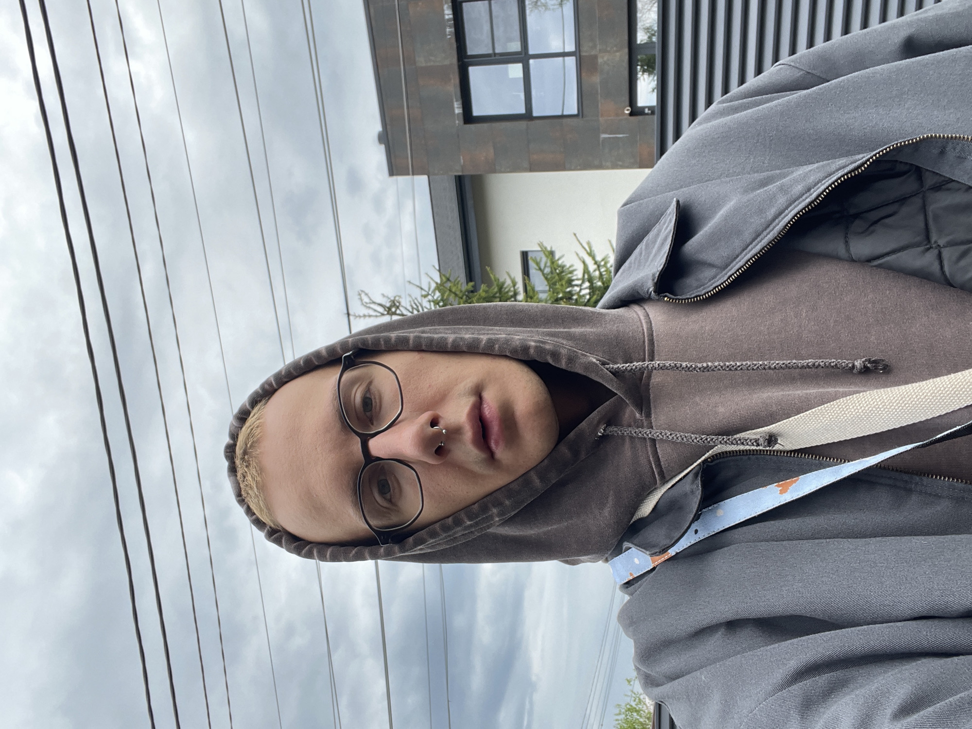 A person wearing glasses, a brown hoodie, and a gray jacket is posing for a picture. They have a nose ring and short blonde hair. In the background, there is a modern building with a black-framed window, and overhead power lines are visible against a cloudy sky. The location is likely in a residential area but a specific location in Sestroretsk, Russia, cannot be definitively determined from the image.