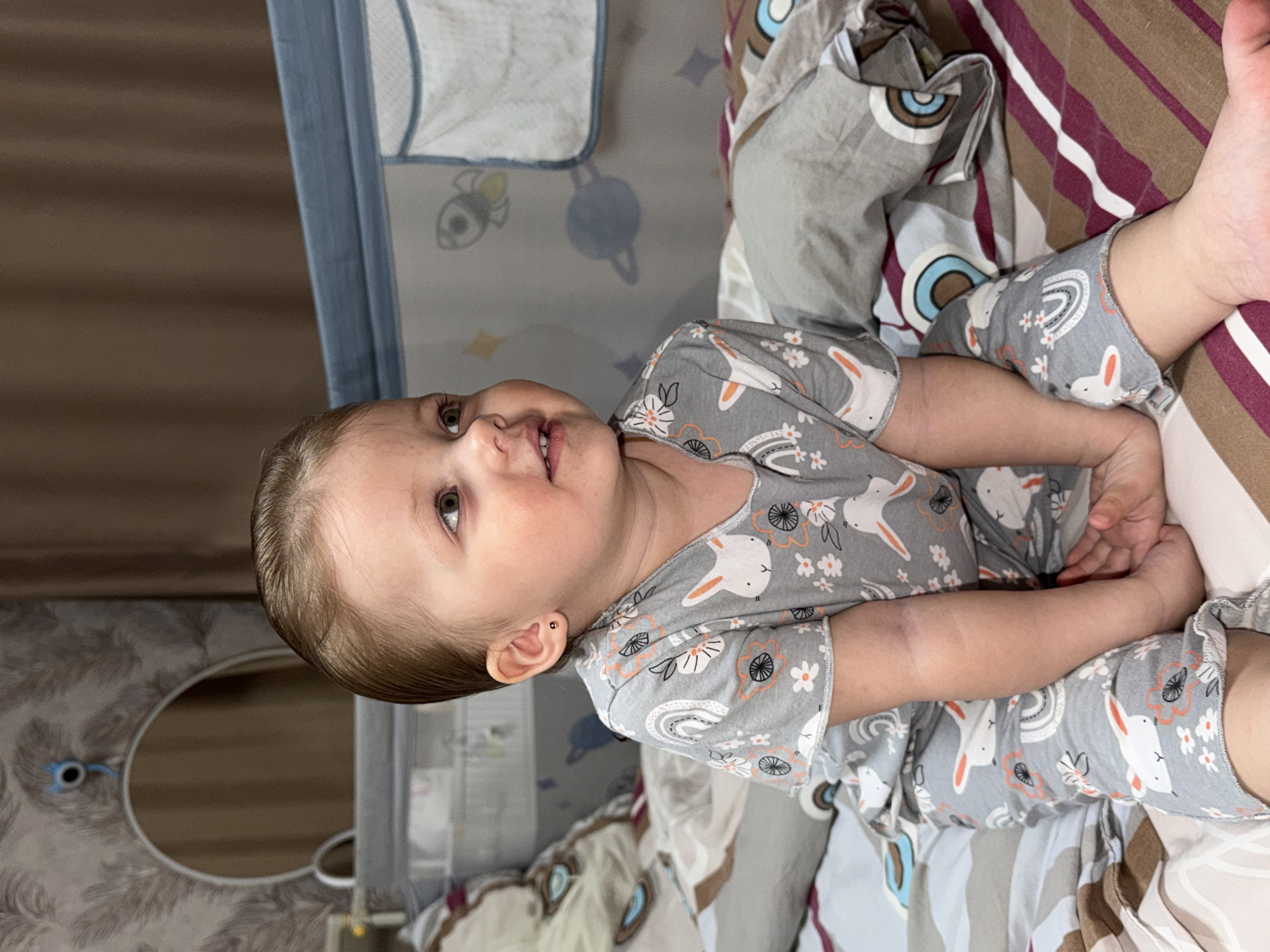 A young child lies in a bed, possibly a crib, in what appears to be a home interior. The child wears gray pajamas patterned with rabbits, rainbows, and flowers. The bedding consists of multiple layers or patterned fabric in shades of gray, brown, maroon, and white. The child’s hands are clasped together in front. A baby crib is visible to the left. The precise location in Reshetnikovo, Russia, cannot be determined based on the provided image.