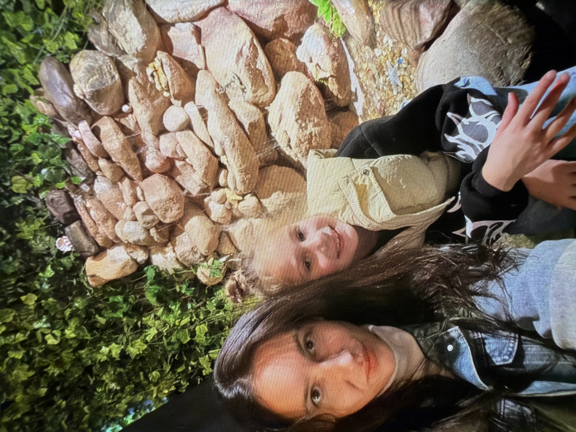 A selfie features two females in front of a rocky background with artificial greenery. The female in the foreground on the left has long, dark hair and is wearing a denim jacket over a cream turtleneck. The female to her right, who appears to be younger, has her hair in a bun and is wearing a light-colored puffer vest over a black sweatshirt with a white graphic design. She is smiling at the camera. The rocky background suggests an indoor exhibit or attraction, possibly a man-made waterfall or grotto. The lighting is artificial, creating a slightly warm tone. The setting is likely an entertainment venue within the Папярнянскі сельскі Савет, Belarus, perhaps a park or a dedicated attraction.