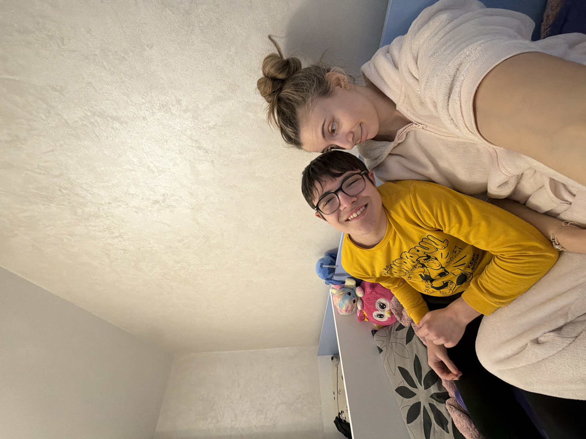 The image displays two individuals seated indoors. A younger person, wearing a yellow long-sleeved shirt with a graphic print and black-rimmed glasses, smiles broadly while leaning their head towards an adult female. The female, with light brown hair styled in a bun and wearing a light pink, fuzzy hooded zip-up garment, looks towards the younger person with a subtle smile, extending her arm to capture a selfie-style photograph.
They are situated on what appears to be a bed or sofa, featuring a light blue frame or headboard. Grey bedding with a dark floral or leaf pattern is visible beneath the younger person. Several stuffed animals are positioned behind the younger person, including a pink owl and a blue character (resembling Stitch). The room's walls are light beige with a distinct textured finish, and the ceiling is plain. No specific architectural or environmental details are present within the image to ascertain the precise location within Plovdiv, Bulgaria.