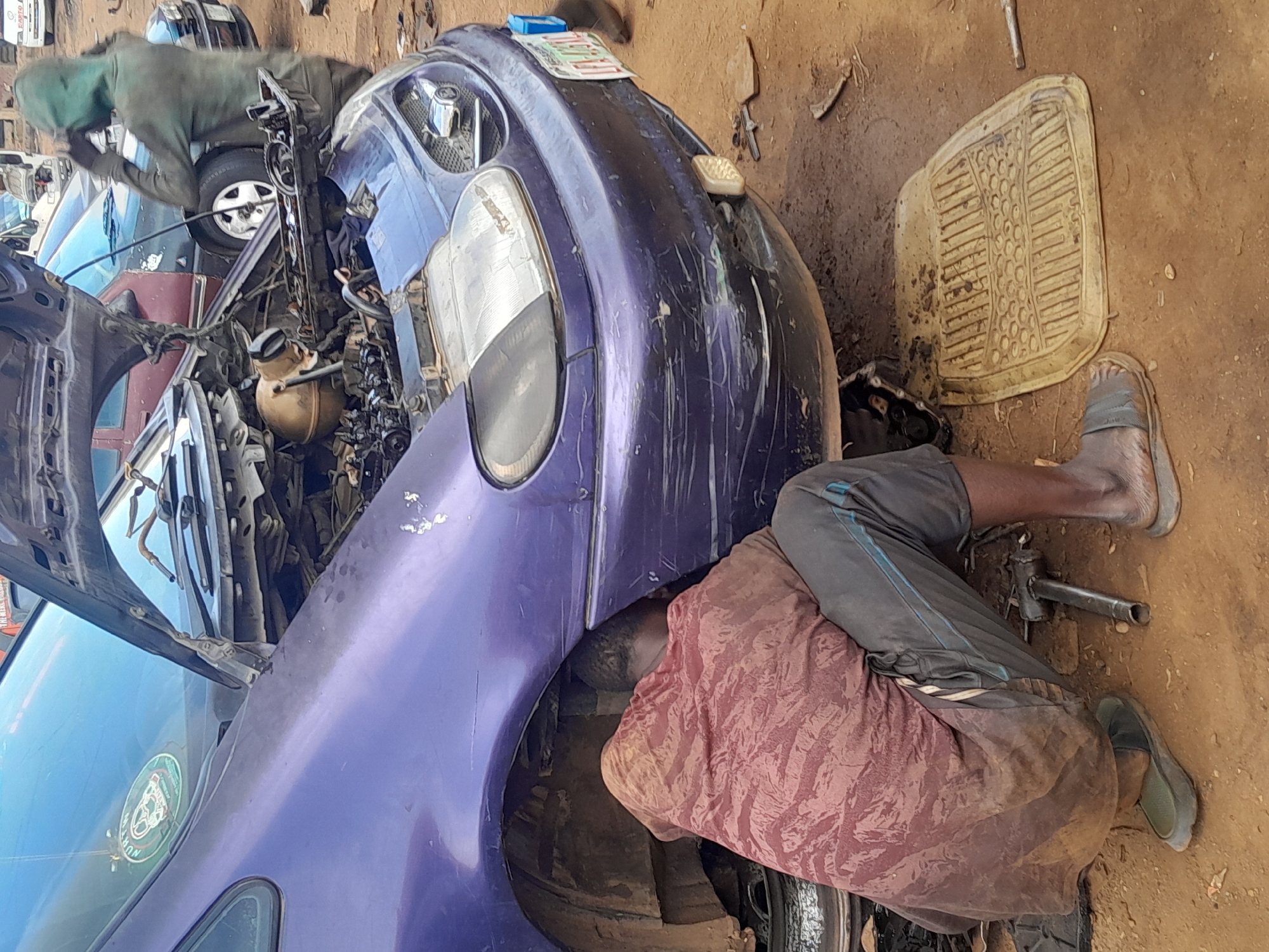 In Jos, Nigeria, an outdoor scene depicts two individuals working on automobiles. One person, wearing a green hooded top, is positioned near the front of a dark red car. A second individual is lying under a purple car. The hood of the purple car is open, revealing the engine compartment. Tools and a rubber car mat are on the ground. Other vehicles and car parts are scattered around the scene.