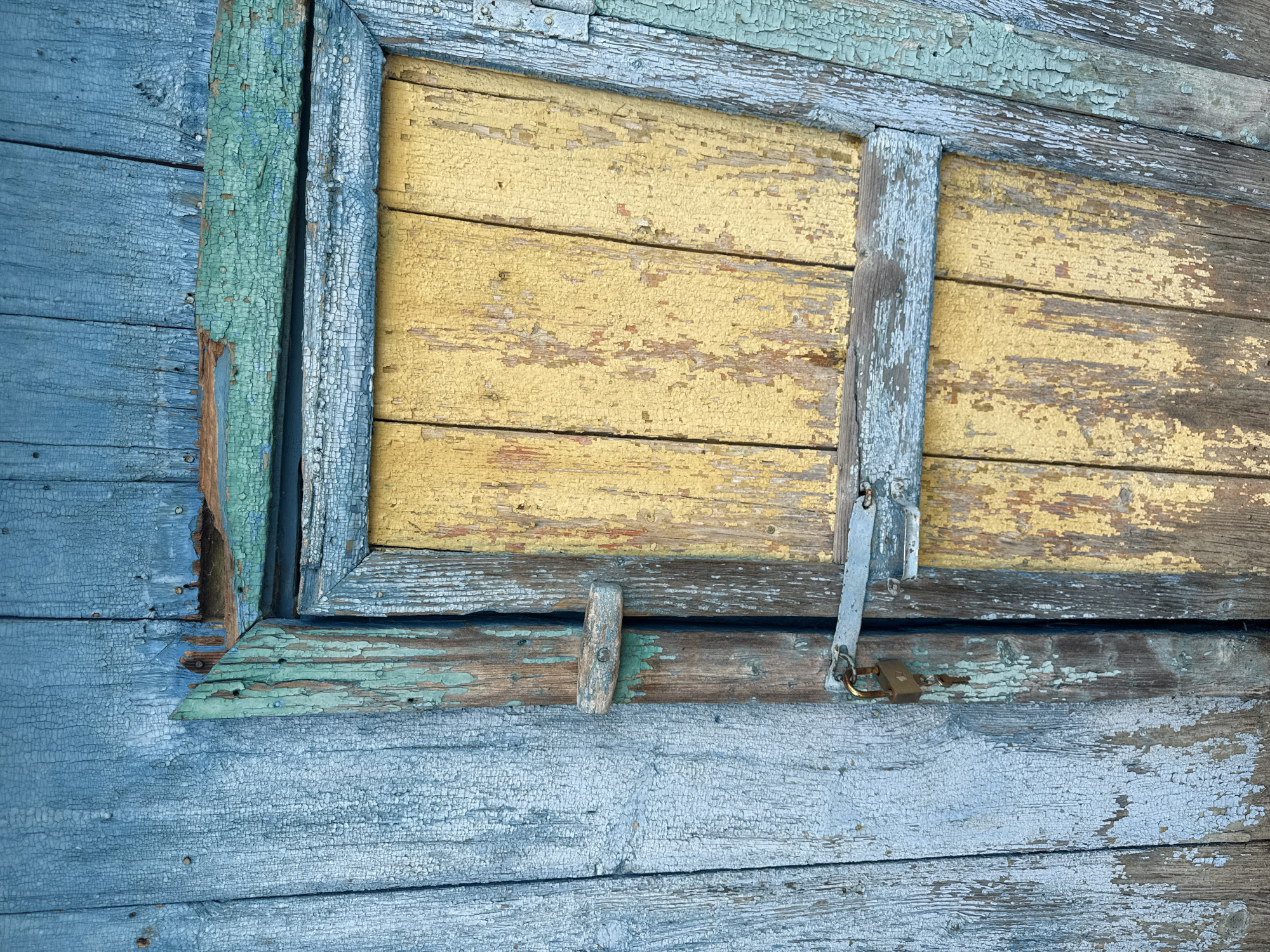 The image shows a close-up of a weathered, blue wooden structure with a small, yellow wooden door. The paint on both the structure and the door is heavily cracked and peeling, revealing the wood underneath. A rusty metal latch is attached to the blue structure, and a padlock with keys hangs from it, securing the yellow door. The wood appears aged and worn, suggesting an outdoor setting, possibly in a rural or industrial area. The overall impression is one of disuse and decay. The location is identified as Чуй-Атасево, Russia.