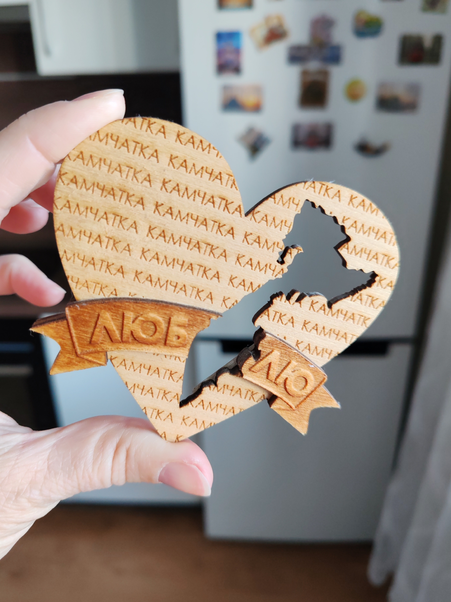 A close-up, eye-level shot captures a person's hand holding a wooden heart-shaped decorative item. The object is crafted from light-colored wood, exhibiting a distinct laser-cut design with slightly darkened, burnt edges. The heart is uniquely split into two jagged pieces, which fit together to form the complete shape. The split line appears to mimic a geographical outline, possibly that of the Kamchatka Peninsula.
The surface of the wooden heart is intricately engraved with repetitive text. The most prominent word, repeated in multiple horizontal rows, is "КАМЧАТКА" (Kamchatka) in Cyrillic capital letters. Towards the bottom, two banner-like elements are integrated into the design, one on each half of the split heart. Together, these banners spell out "ЛЮБОВЬ" (Lyubov), meaning "Love" in Russian, with "ЛЮБ" on the left piece and "ОВЬ" on the right.
The background is softly blurred, revealing a white refrigerator covered with a collection of small, colorful magnets and printed photographs, suggesting a personal and homely indoor setting. The scene is illuminated by bright, natural light, indicative of daytime. The item appears to be a souvenir or a decorative piece commemorating the Kamchatka region, held up for display in what seems to be a residential kitchen or living space.