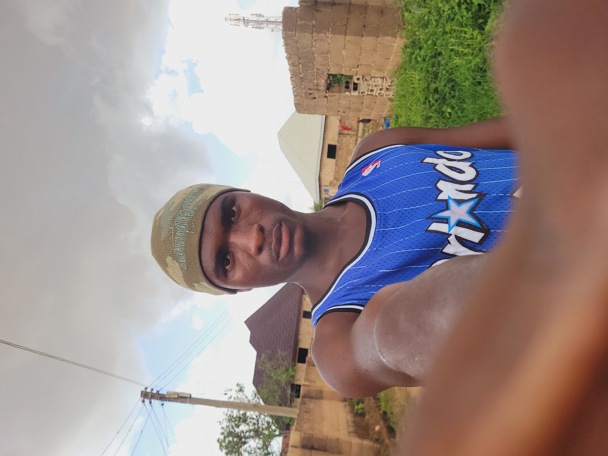 A medium-shot, eye-level, selfie-style photograph shows a young Black male with dark skin, wearing a green knit cap and a blue basketball jersey with white pinstripes and "Orlando" written across the chest in white lettering with a blue star. He is looking directly at the camera with a neutral expression. His right arm is extended towards the camera, with his hand and thumb out of focus in the foreground, obscuring a portion of the image.
The background suggests an outdoor, semi-urban environment in Bukuru, Nigeria. To the left, a utility pole with electrical wires is visible. Behind the subject, a low, tan-colored brick wall runs horizontally. Beyond the wall, there are buildings under construction, one with a brown, pitched roof and another with a white, triangular roof. To the far right, a partially constructed building with an open doorway made of rough-hewn tan bricks stands amidst green grass and foliage. A tall telecommunications tower is visible in the distance above this structure. The sky is overcast with white and grey clouds. The lighting suggests it is daytime.