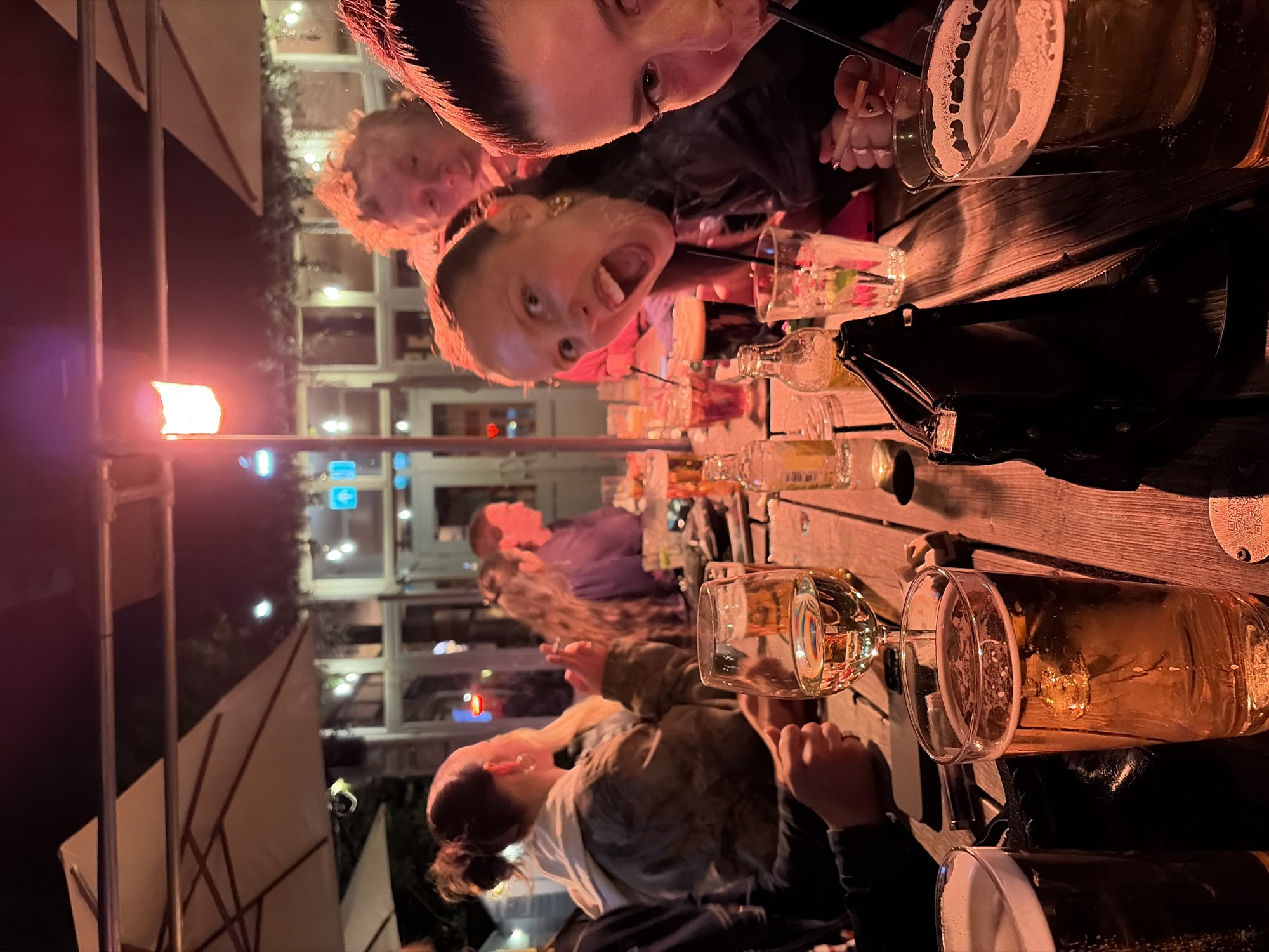The image shows a group of people at an outdoor table, possibly at a pub or bar, in London, United Kingdom. Many drinks are on the table, including beers and cocktails, and the people appear to be socializing and enjoying themselves. The table is made of wood. The lighting suggests it is nighttime, with artificial lights illuminating the scene. The background includes architectural details typical of London, with multi-story buildings and outdoor seating areas visible.
