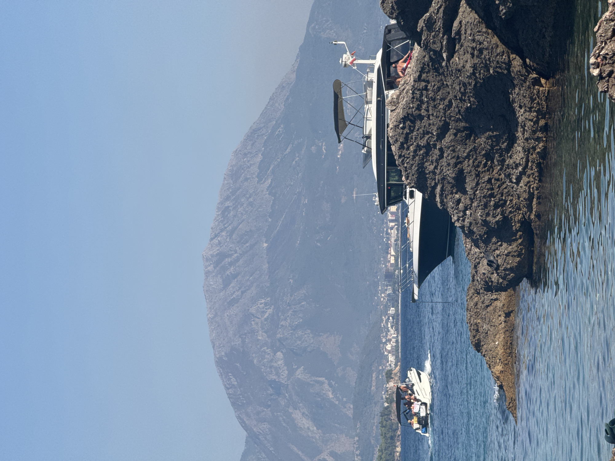 In Sutomore, Montenegro, two boats navigate the azure waters of the Adriatic Sea, with a majestic, mist-shrouded mountain range serving as a dramatic backdrop. In the foreground, dark, rocky outcrops jut out from the water, creating a natural, rugged border. The boat on the right, a sleek, modern vessel, is anchored near the rocks, its dark hull contrasting with the bright sky. A few individuals are visible lounging on its deck. To the left, a smaller, white speed boat cuts through the waves, carrying a group of people enjoying a ride. A coastal town, with its white buildings and red-tiled roofs, is nestled at the base of the mountains, further accentuating the serene beauty of the scene. The sky above is a clear, unblemished blue, hinting at a warm, sunny day.