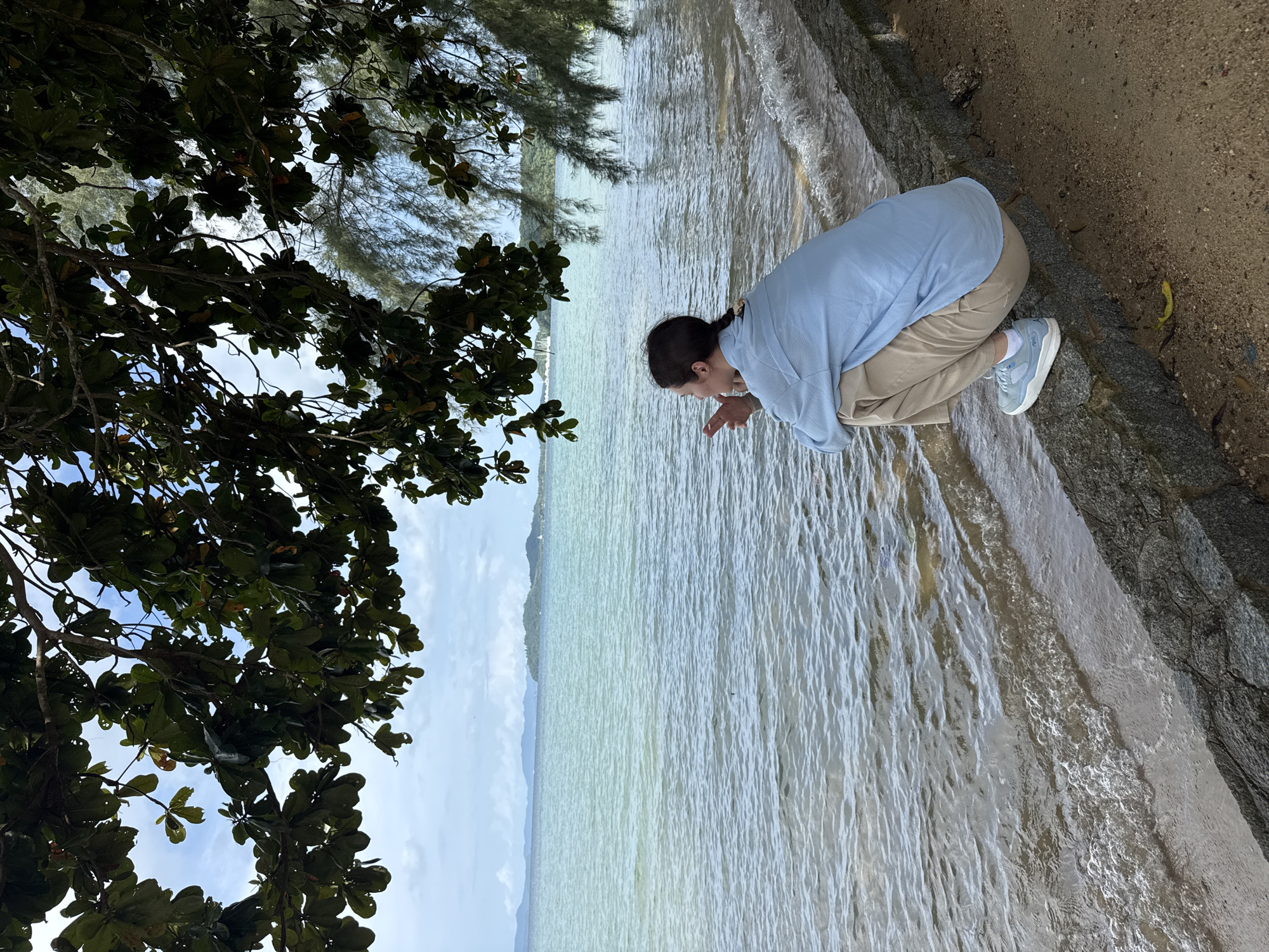 A person kneels on a stone embankment beside the ocean in Pa Khlok, Thailand. The person is wearing a light blue button-down shirt, tan pants, and light blue sneakers, and is facing away from the camera, looking out at the water. They have their hands clasped near their chin. The ocean is calm with small waves lapping at the shore. Trees with green leaves hang over the scene from the left and right. In the distance, islands and mountains are visible on the horizon under a cloudy sky.