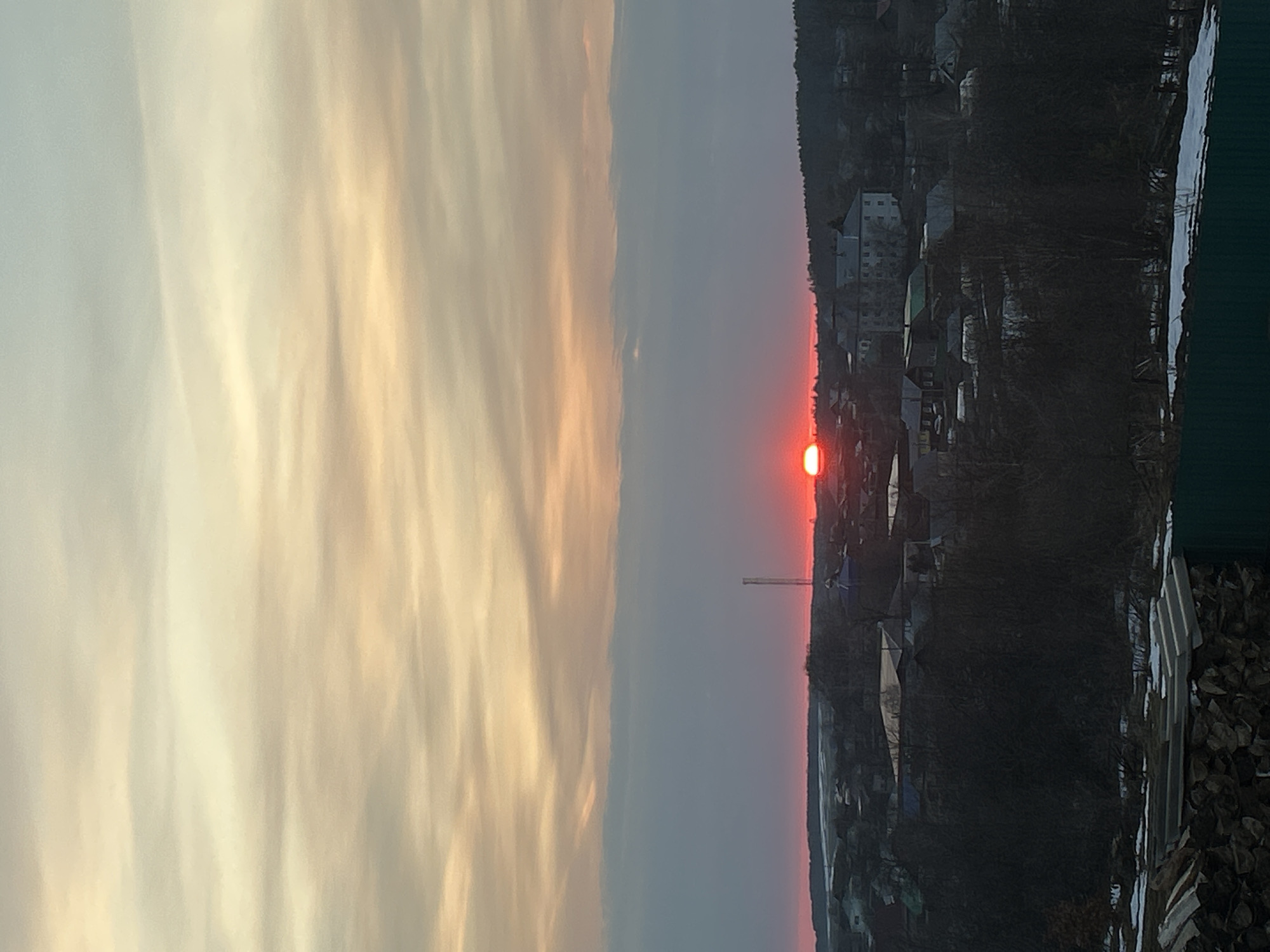 The image shows a sunset over the city of Фоки, Russia. The sky is divided into two distinct sections: a light yellow cloudscape on the left and a gray-blue expanse with the setting sun on the right. The sun appears as a bright red orb just above the horizon. Below the horizon, there is a residential area with buildings and trees. Some snow can be seen on the ground in the foreground. A tall metallic structure (possibly an antenna or a tower) is also visible. The overall scene depicts a winter or late autumn sunset in a small Russian town.