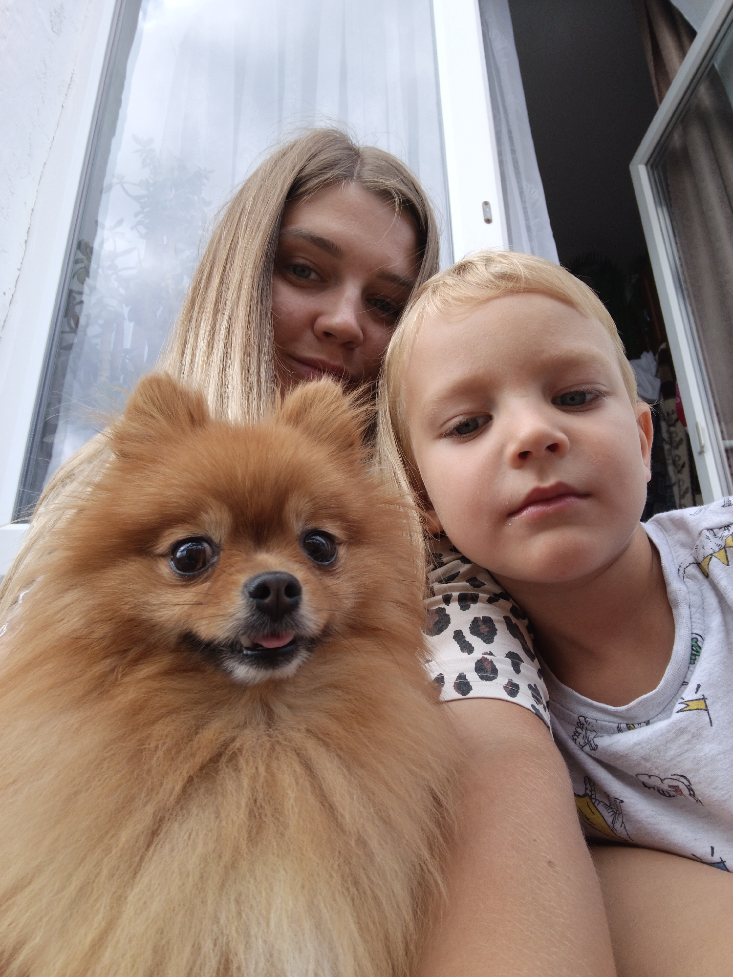 A woman, a young boy, and a Pomeranian dog are posing for a selfie outdoors. The woman has long blonde hair and is smiling. The boy has blonde hair and is looking down. The dog has light brown fur and is looking at the camera with its tongue sticking out slightly. They are positioned in front of a white window frame, with a window showing reflections of trees and sky. The background appears to be a residential setting, possibly a house in the city of Medvedyovka, Russia.