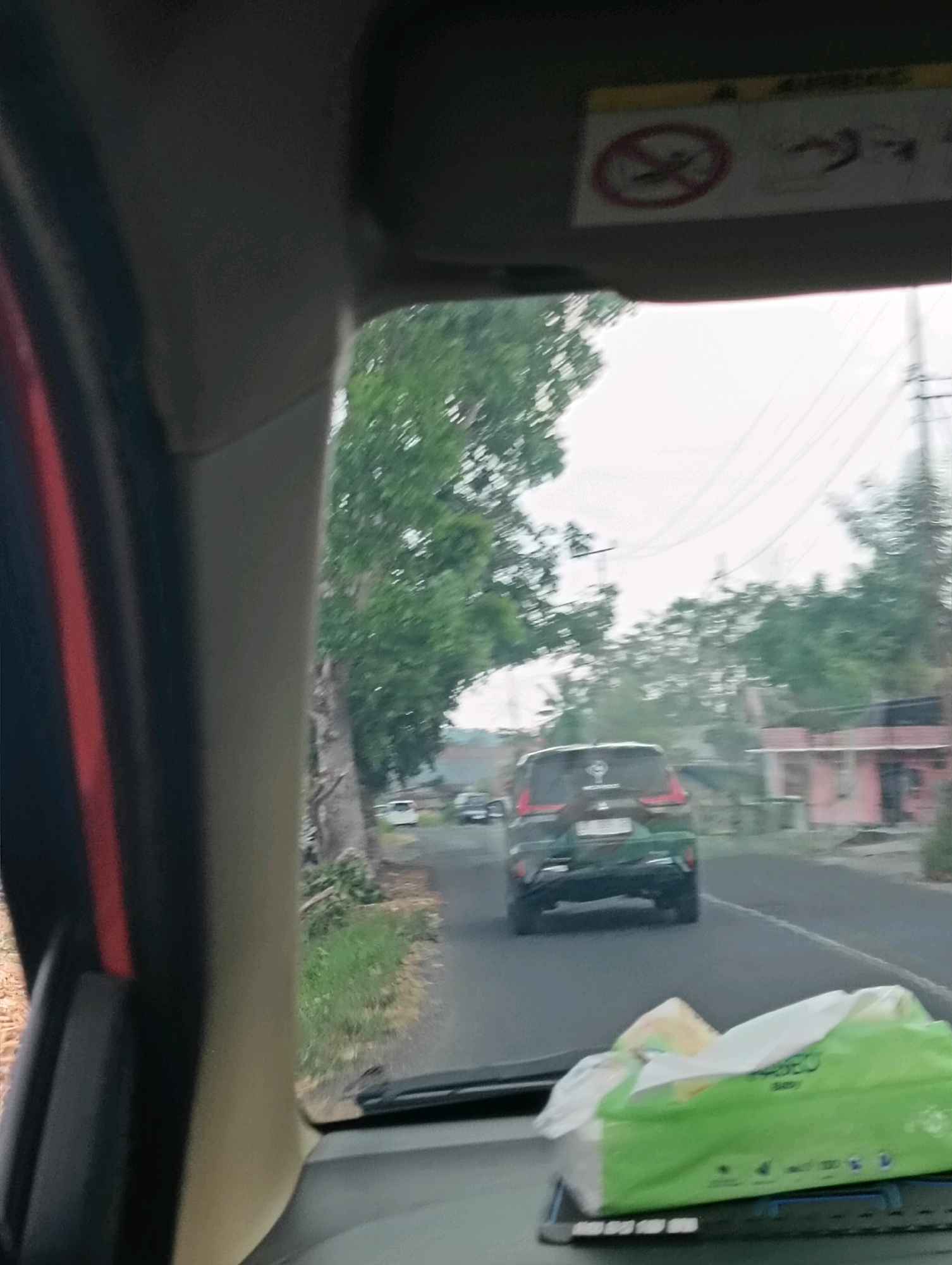 The image is taken from inside a vehicle looking forward, with a dashboard visible in the foreground. A package of green tissue is on the dashboard. The vehicle appears to be on a road in a semi-urban or rural area. Several trees line the left side of the road. In the distance, a dark-colored SUV is driving on the road. Power lines are visible overhead. On the right side of the road, there are buildings with pink accents. The sky is overcast, suggesting it might be a cloudy day. The image does not provide specific visual cues to confirm the location as Tomohon, Indonesia, but the general scenery of lush greenery and residential buildings could be consistent with that region.