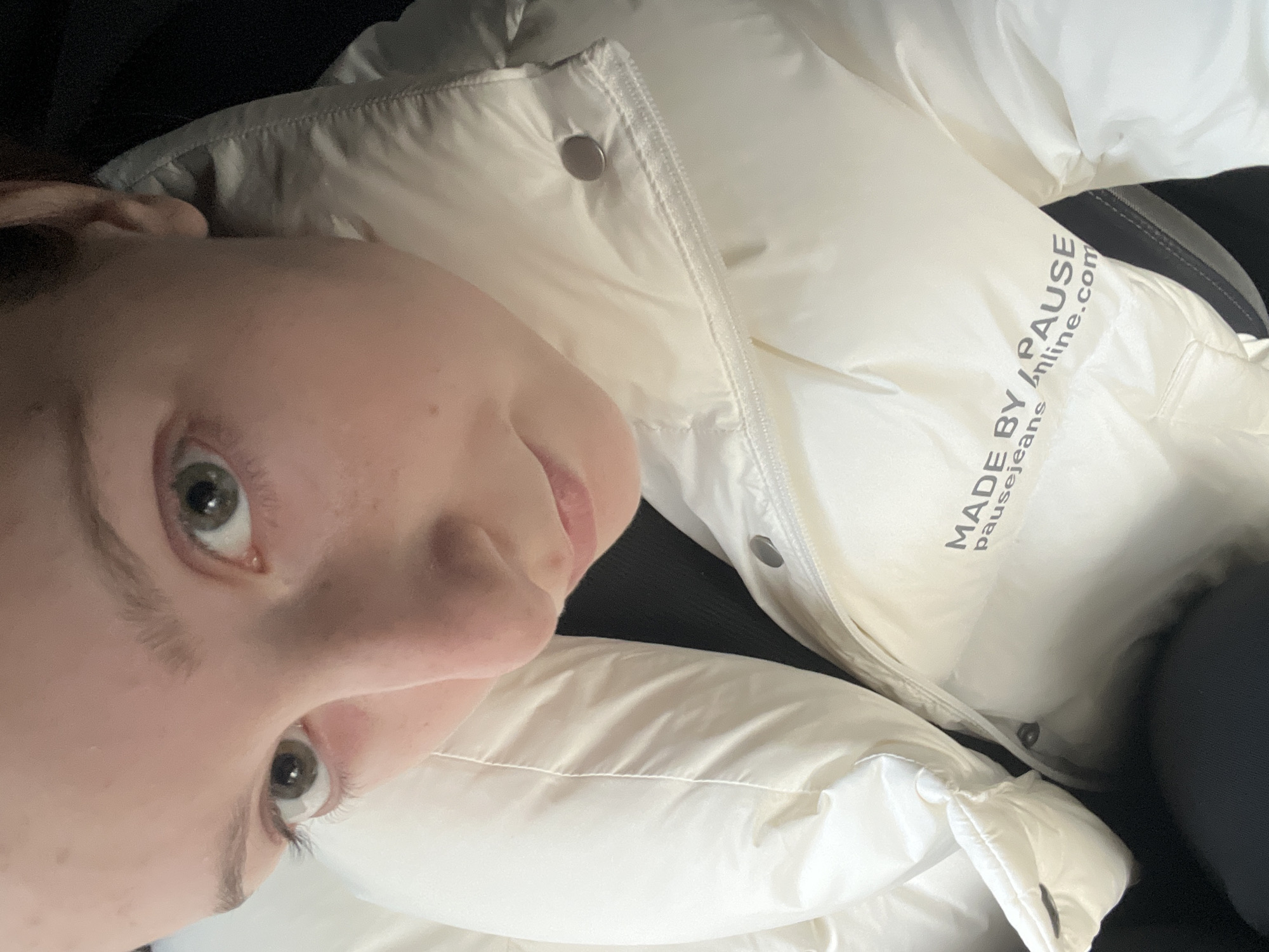 A close-up, slightly tilted selfie shows a person with fair skin, light green eyes, and indistinct hair color. They are wearing a puffy, off-white jacket with metallic snap closures along the zipper. The jacket's fabric appears smooth and somewhat reflective. The jacket features a printed logo "MADE BY / PAUSE pausejeansonline.com". The background is predominantly dark, suggesting an indoor setting or low lighting conditions. There's no indication of Varna, Bulgaria in the image.