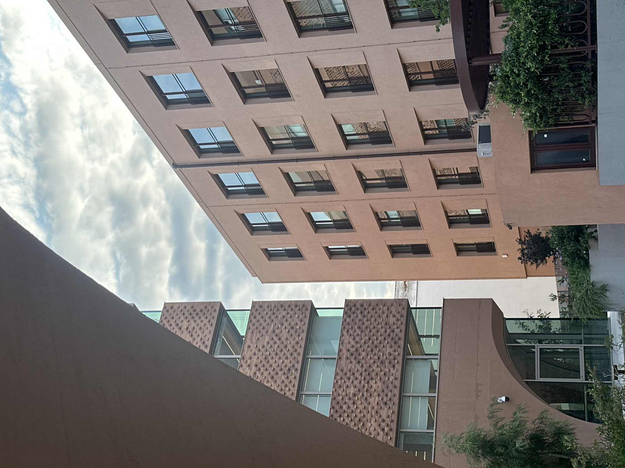 The image shows the exterior of two modern buildings in Thessaloniki, Greece. The building on the left features a distinct facade with textured brickwork and large glass windows. The building on the right is a light pink, multi-story structure with numerous identical windows, each with a small black balcony railing. The sky above is partly cloudy with a soft, diffused light, suggesting it might be early morning or late afternoon. Greenery, including trees and bushes, is visible in the foreground and around the base of the buildings, indicating an urban setting with landscaping. There are no people visible in the image.