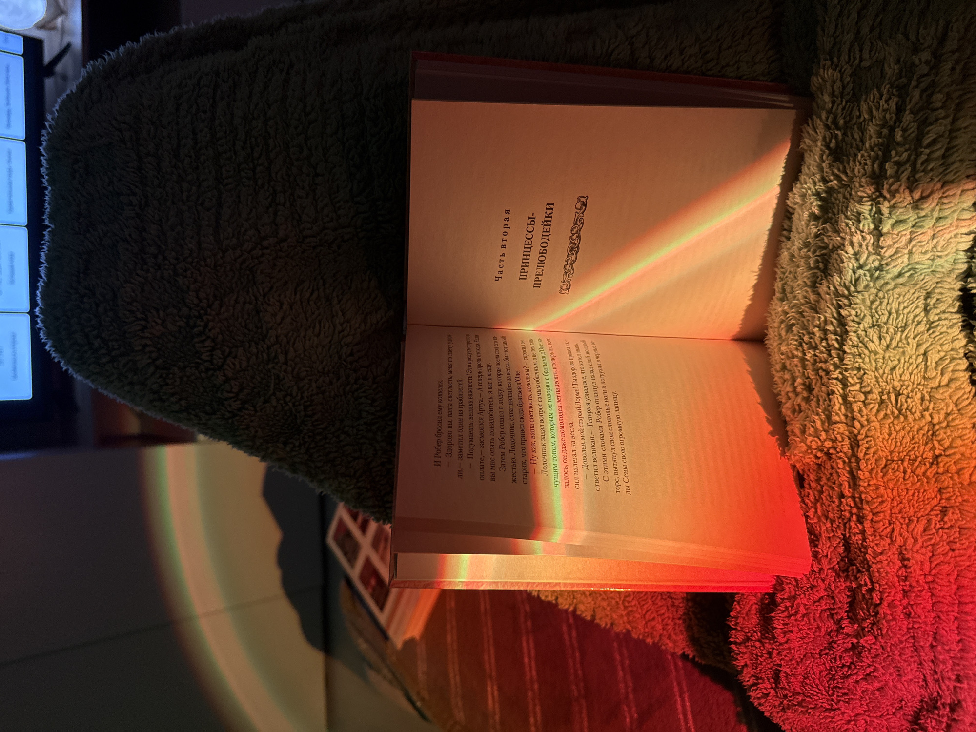 An open book rests on a textured, greenish-brown fuzzy blanket, illuminated by a striking artificial light source that casts vibrant rainbow-like streaks across the pages and the surrounding fabric. The scene appears cozy, likely taking place in the evening or at night due to the prominent warm glow and deep shadows.
The book is a hardcover with off-white pages. The visible text is in Russian. The right page displays a chapter title: "ЧАСТЬ ВТОРАЯ" (PART TWO) and below it, "ПРИНЦЕССЫ-ПРЕЛЮБОДЕЙКИ" (PRINCESSES-DEBAUCHERS/PROSTITUTES). The body text describes a narrative involving characters like Robert, Artois, and an old boatman named Lorme, with mentions of money, a boat, and the Seine river. Phrases such as "И Робер бросил ему кошелек" (And Robert threw him a purse) and "Доволен, мой старый Лорме!" (Satisfied, my old Lorme!) are visible.
The blanket's shaggy texture is emphasized by the light and shadows. To the far left in the background, a glowing, out-of-focus blue screen or digital display can be seen. The overall mood is intimate and relaxed, suggesting an act of reading or leisure under a unique ambient light.