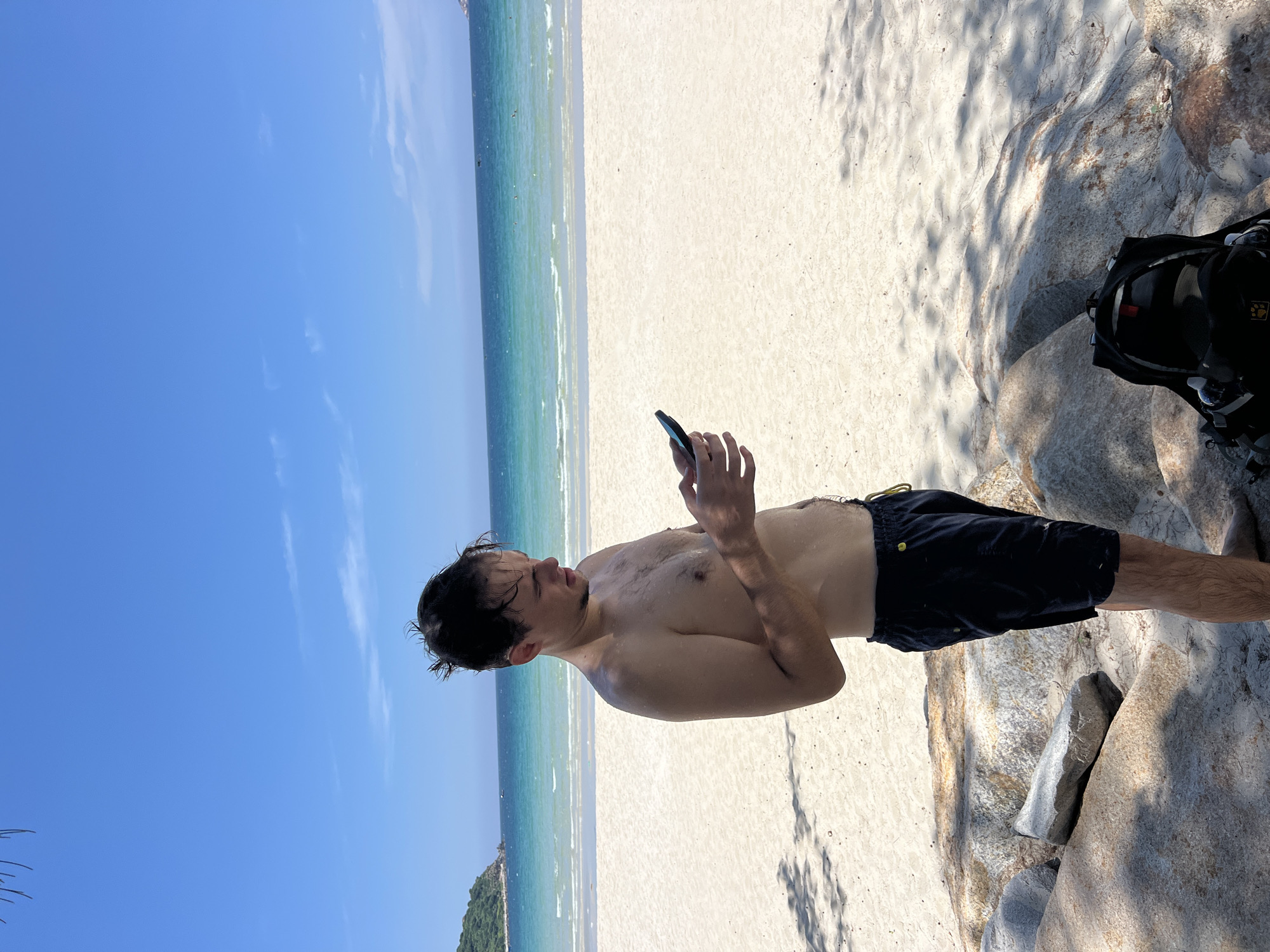 A shirtless young man stands on a sandy beach in Cong Hai, Vietnam, looking at his cell phone. He is wearing dark blue swim trunks. The beach is white sand, and the ocean is turquoise and blue with gentle waves. There are some rocks and a black backpack on the sand in the foreground. In the background, there is a green, hilly island. The sky is bright blue with a few scattered clouds. The man appears to be wet, possibly from swimming. The scene is lit by bright sunlight, casting shadows from nearby trees.