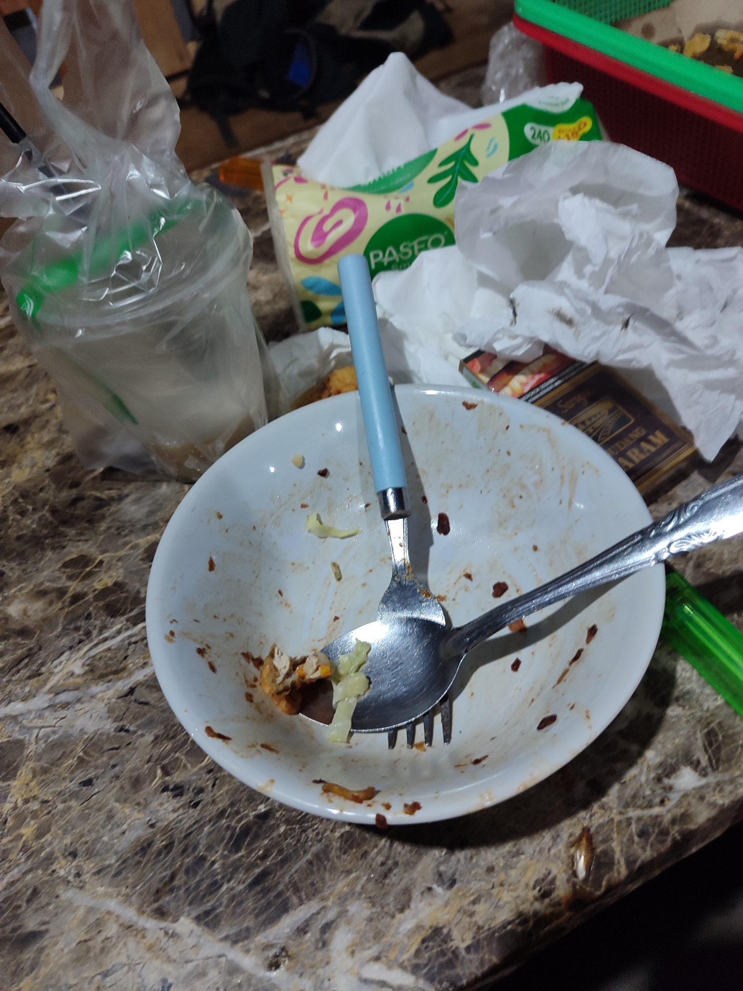 The image depicts a close-up, overhead view of a dirty white bowl with leftover food, a fork, and a spoon on a marble-patterned table. To the left, a clear plastic bag contains a drink with a green straw. Behind the bowl, there is a crumpled tissue and a partially visible pack of cigarettes. A green plastic object is also present in the lower right corner. The scene suggests a meal has recently concluded. The location is identified as Gantungan, Indonesia, though no specific landmarks or environmental context are visible to confirm this.