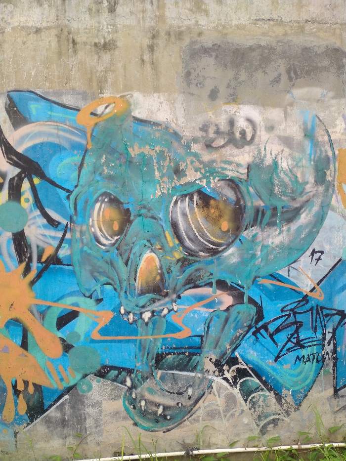 This media file depicts a vibrant and weathered graffiti mural on a concrete wall, likely outdoors in Karanganyar, Indonesia. The central subject of the graffiti is a large, stylized skull or monstrous head, predominantly rendered in shades of blue and teal. Its surface shows significant texture and areas where the paint appears chipped or worn, either intentionally or due to age.
The skull features large, expressive eyes with glowing orange-yellow centers, highlighted by black and white details. An orange, halo-like ring with drips sits above the skull's head, and splashes of orange paint are scattered on the left side of the artwork, contrasting with the blue background. The graffiti background itself consists of various shades of blue with black and white outlines and abstract shapes.
The concrete wall above and around the graffiti is visible, showing signs of aging, with weathered grey and beige patches. At the very bottom of the image, green grass and a white pipe are discernible, indicating the wall is at ground level.
Visible text within the graffiti includes the number "17" on the right side of the skull. Below this, towards the bottom right, there is stylized black tagging, with the word "MATURA" clearly legible underneath. The image appears to be taken during daylight.