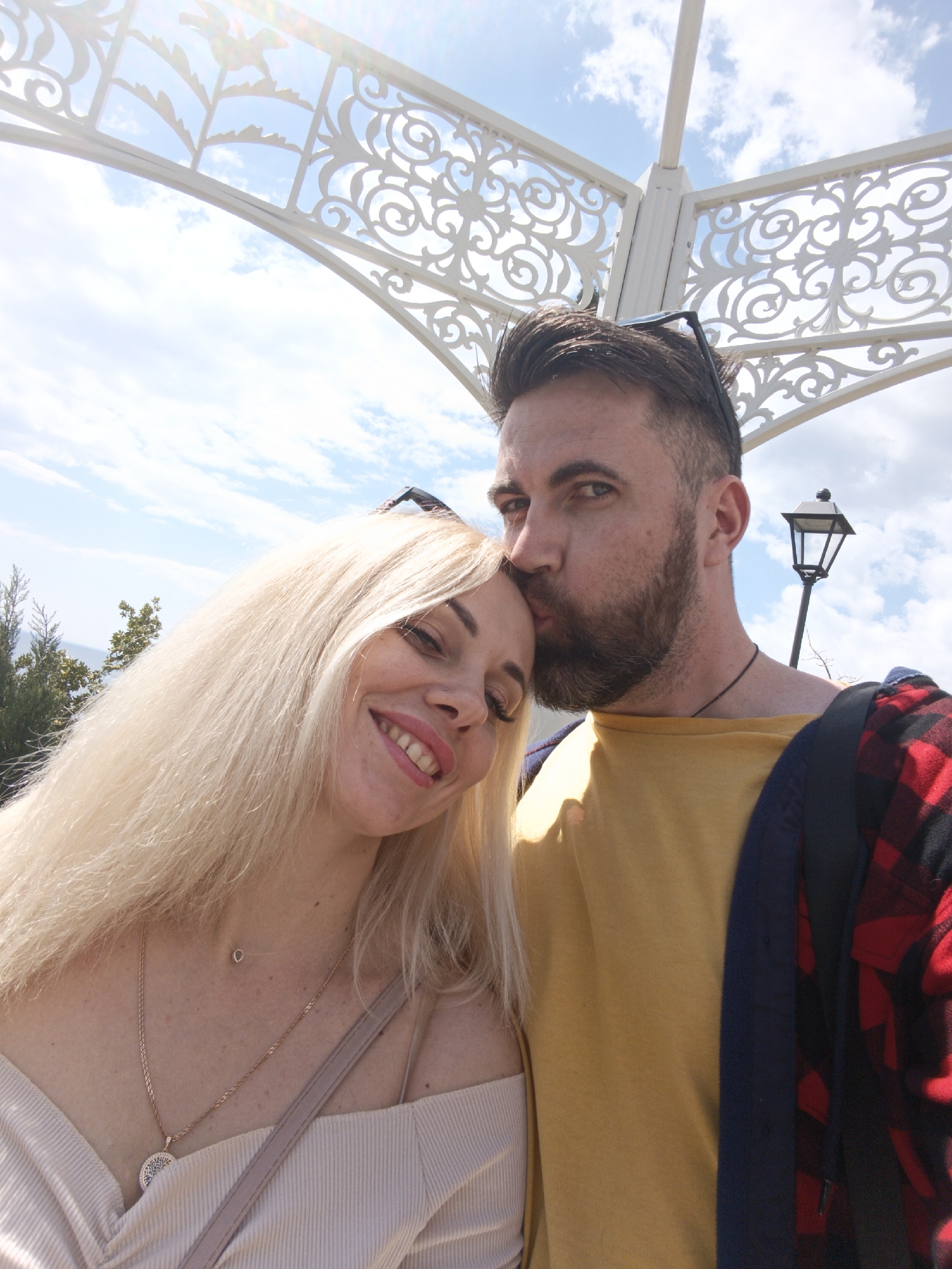 A selfie shows a man kissing a woman on the forehead. The man has dark hair, a beard, and is wearing a yellow shirt with a red and black flannel shirt draped over his shoulder. The woman has blonde hair and is wearing a light-colored top. They are standing under a white, decorative archway. In the background, there is a street lamp, trees, and a blue sky with clouds. The location is in the city of Unknown, Unknown.