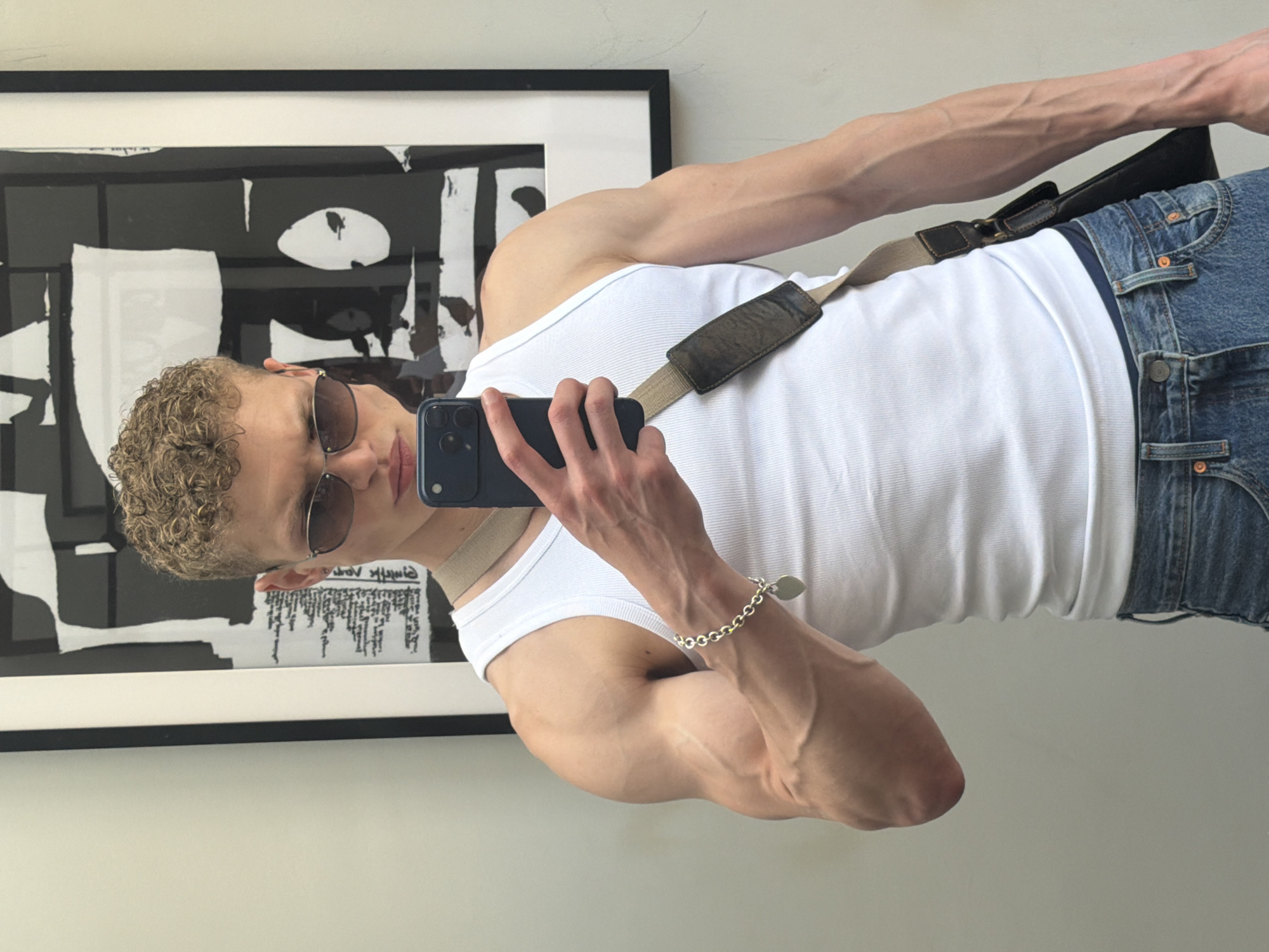 A mirror selfie taken indoors depicts a young person with short, curly, light brown hair and sunglasses, holding a dark-colored smartphone to capture their reflection. The person is positioned in a casual, standing pose, looking into the phone's screen. They are wearing a white ribbed tank top and blue denim jeans. A brown/khaki-colored strap, possibly from a bag, crosses their right shoulder. A silver chain bracelet with a small, light-colored charm is visible on their left wrist. Their arms display a lean, muscular build with prominent veins.
The background consists of a plain, light-colored wall. Directly behind the person's head and upper body, a framed piece of abstract artwork hangs on the wall. The artwork is predominantly black and white, featuring bold, geometric, and organic shapes. Within a white section of the artwork, some stylized, reversed text is visible, although it is unreadable due to the angle and artistic representation. The lighting is bright and diffused, suggesting a daytime setting. The scene appears to be a modern indoor environment in London, United Kingdom.