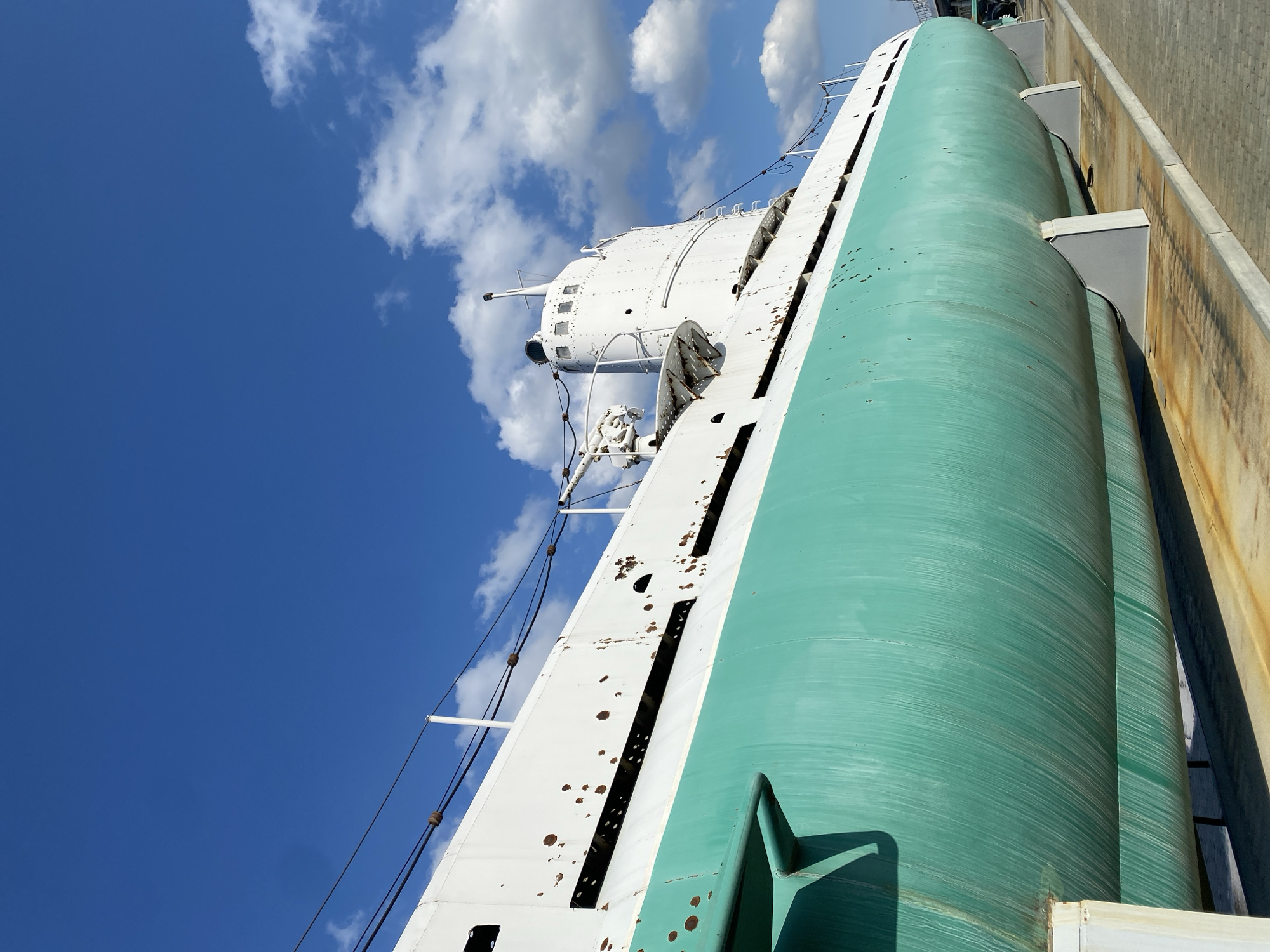 The image shows a decommissioned submarine, painted in white and teal, resting on a display stand in the city of Verkhnyaya Pyshma, Russia. The submarine is positioned outdoors under a clear blue sky with scattered white clouds. The hull of the submarine exhibits signs of wear, with rust spots visible on the white upper section. A periscope and other external structures are mounted on the conning tower. The submarine appears to be part of a museum or memorial, as it is supported by sturdy grey stands and situated on a paved area, possibly an exhibition ground. The background is dominated by the expansive sky, with hints of greenery and other structures visible in the far distance, suggesting an outdoor, possibly urban or industrial, setting.