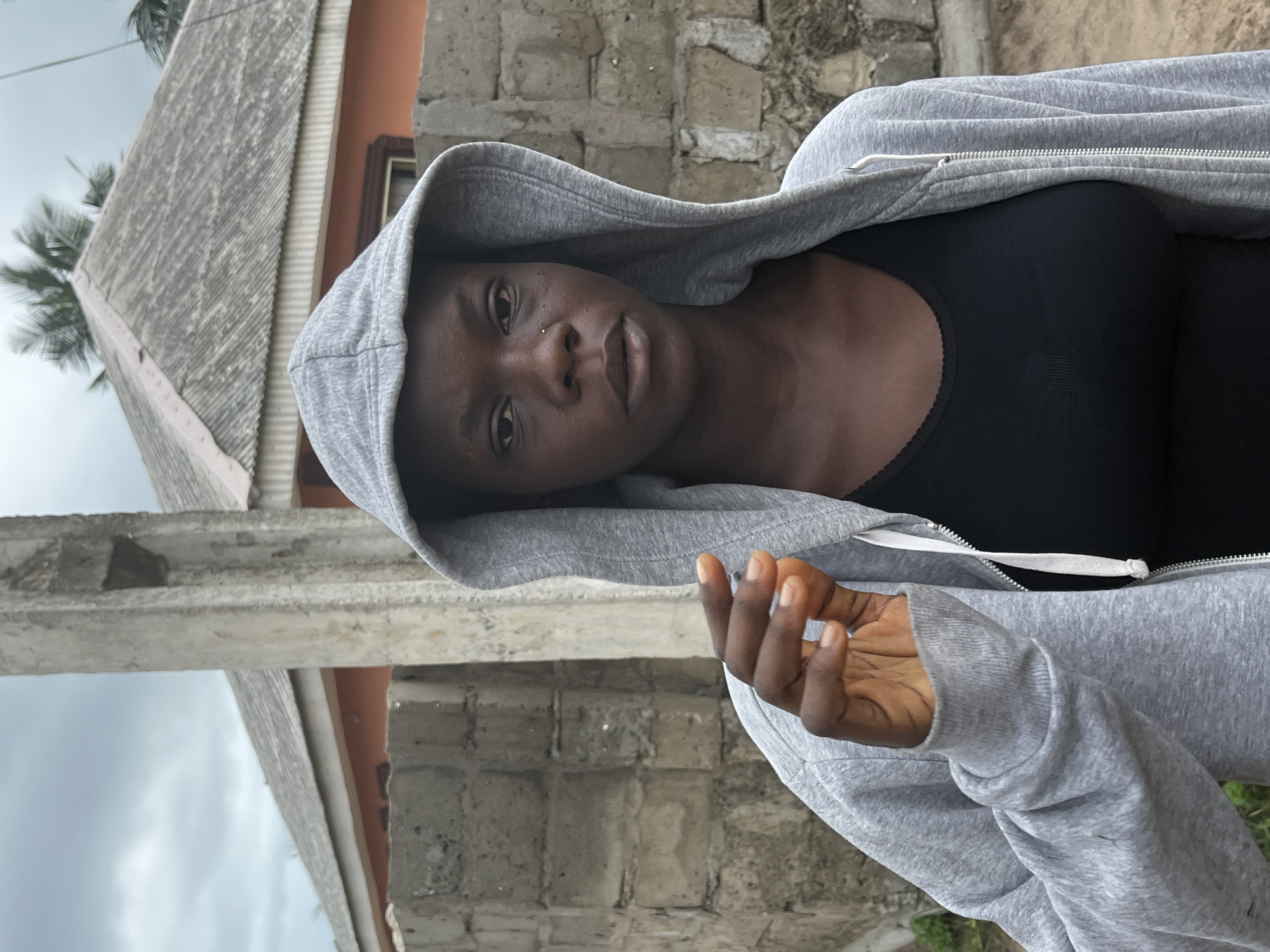 A dark-skinned person with a nose piercing is wearing a gray hooded sweatshirt over a black top. They are standing outdoors in front of a building, possibly in Badagry, Nigeria. The building appears to have a concrete block wall and a gray roof. Part of the sky is visible.