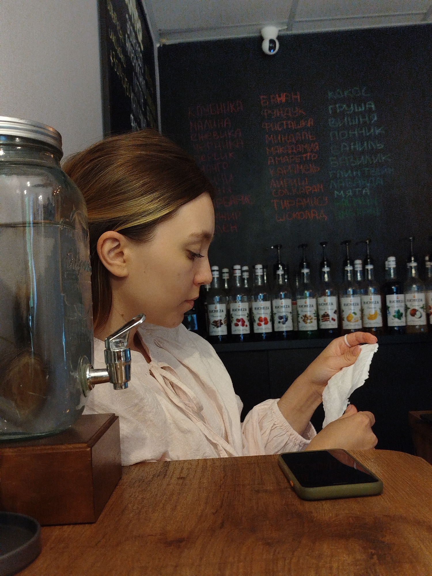A young woman with blonde hair pulled back is seen in profile, looking down at a white paper napkin she is holding in her hands. She is wearing a light pink, long-sleeved blouse. To her left is a large glass dispenser filled with a clear liquid and a silver spigot. The dispenser sits on a dark wooden box. Behind her, a blackboard lists various items in colorful chalk lettering, likely menu items or product names. Below the blackboard, a row of glass bottles with dark liquid and labels is arranged on a shelf. A smartphone with a dark screen and a green case lies on the wooden surface in the foreground. The overall environment appears to be a cafe or bar.