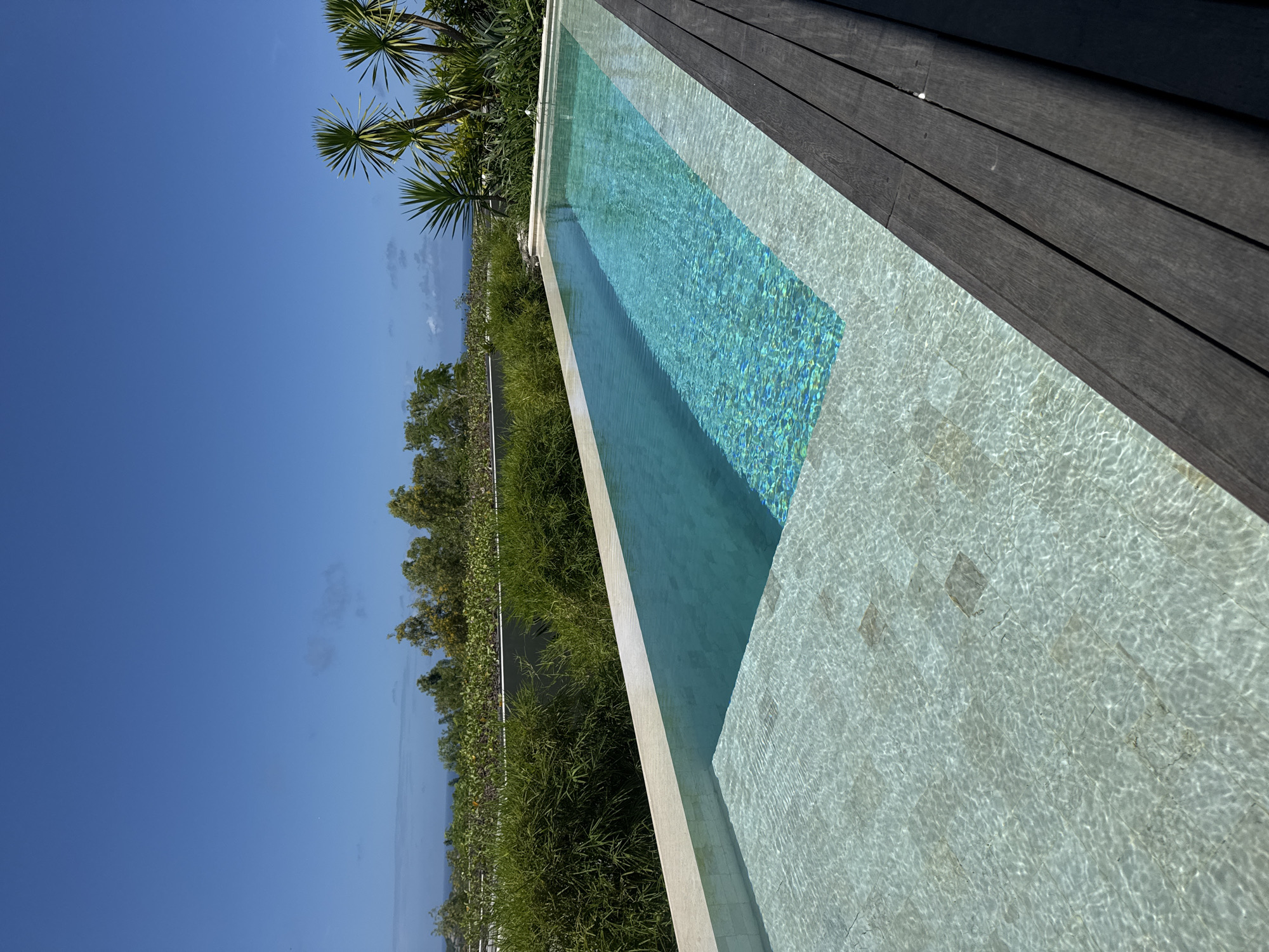 A sideways perspective shows a sleek, narrow swimming pool with varying water depths, accompanied by lush greenery, palm trees, a wooden deck, and a clear, sunny sky. The pool features a deeper turquoise section and a shallower, light-colored area with visible tile patterns under the water's surface. A well-maintained garden with vibrant green plants and palm trees borders one side of the pool, contrasting with the dark brown wooden deck on the opposite side. The clear blue sky suggests a bright, sunny day, typical of a tropical setting. The location is Kuta Selatan, Indonesia, as specified.