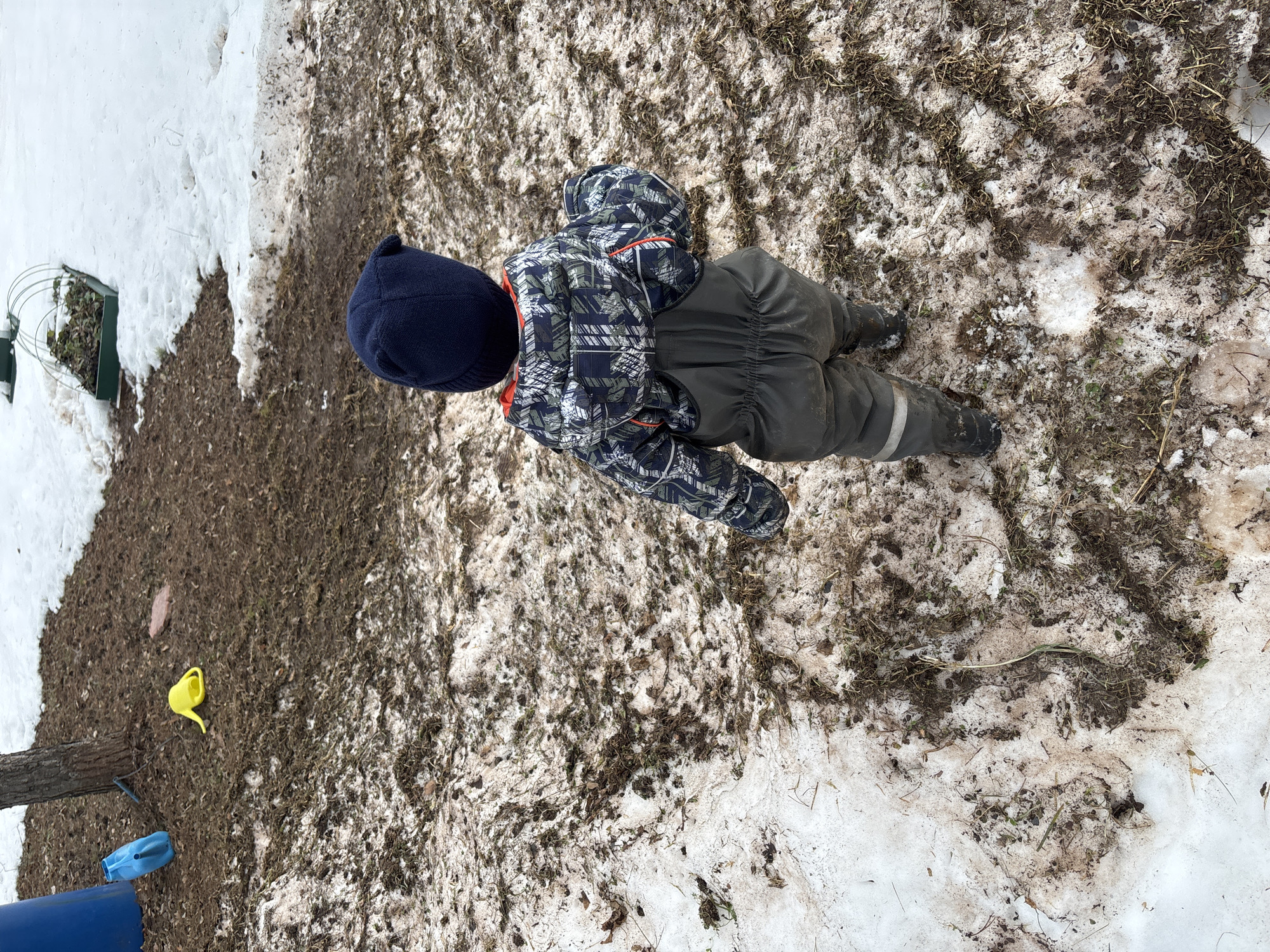 A young child lies prone on a mixture of melting snow and exposed soil, located in Ферма, Russia. The child is wearing a dark blue knit hat, a patterned snow jacket (navy blue, gray and green), and dark gray snow pants with boots. The ground around the child shows a mix of snow, bare soil, and scattered debris such as dry grass. In the periphery, a partially buried garden bed and some water containers can be seen.