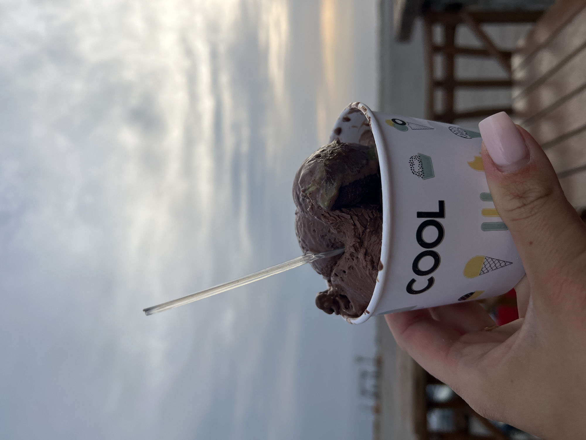 A medium shot depicts a hand holding a cup of chocolate ice cream with a spoon inserted into it. The cup is white with the word "COOL" printed vertically in black, along with other small dessert-themed illustrations. The background features a cloudy sky above what appears to be a shoreline with buildings visible in the distance. The location is not identifiable as the city of Kovačko Polje, Montenegro from the image.