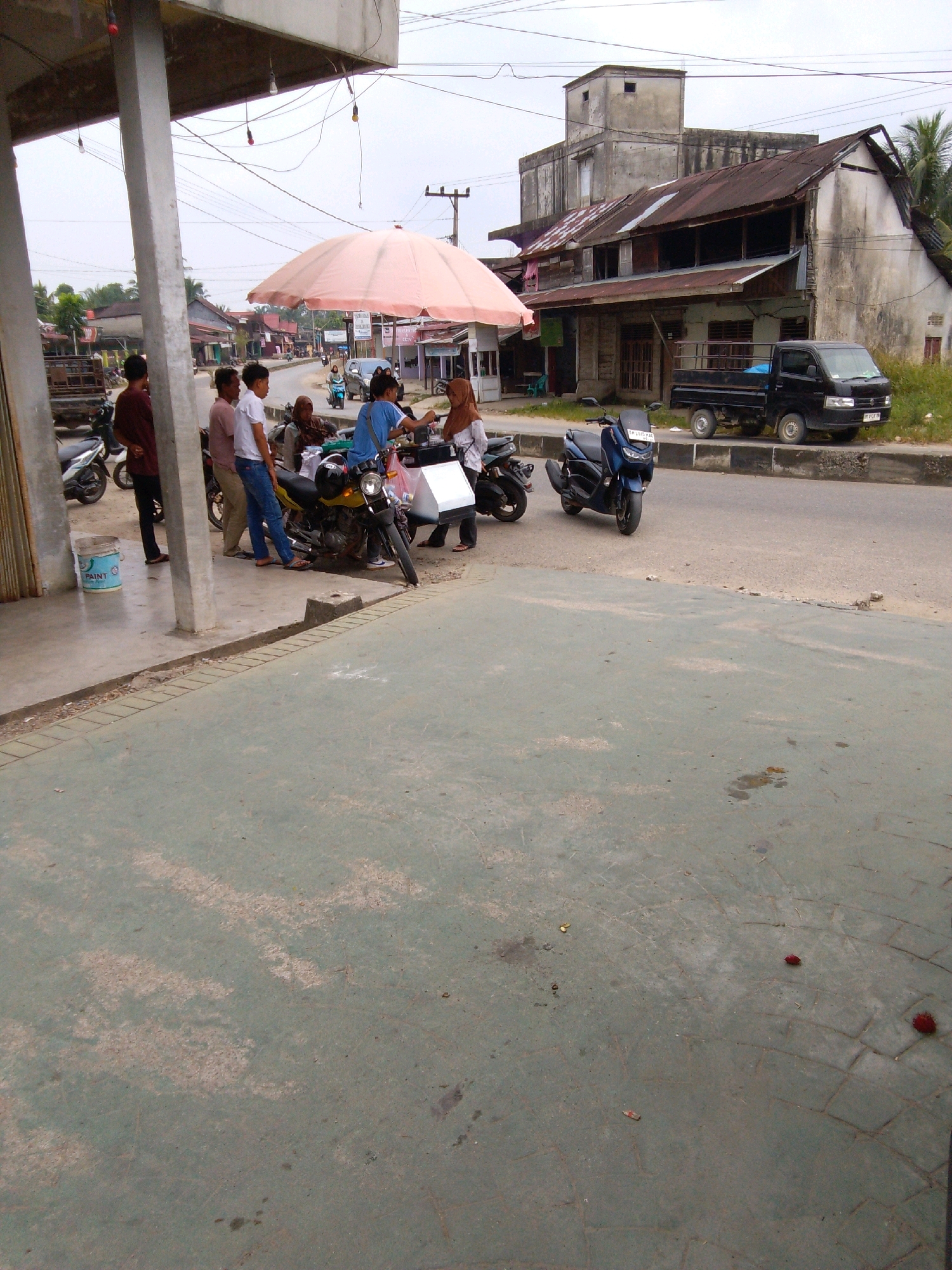In Tangun, Indonesia, a group of people are gathered near motorcycles and a truck parked on a paved road next to a building. A large pink umbrella provides shade over a yellow motorcycle. Several individuals, including men in casual clothing and women in hijabs, are seen interacting. One man, wearing a red shirt, stands by a motorcycle. Another man in a white shirt and blue jeans stands next to him. A woman in a blue shirt is seated on the yellow motorcycle. Adjacent to the motorcycles, a black truck is parked on the side of the road. The background shows a weathered building with a rusty roof, and beyond that, a street lined with more buildings. The ground in the foreground is a light gray concrete surface.