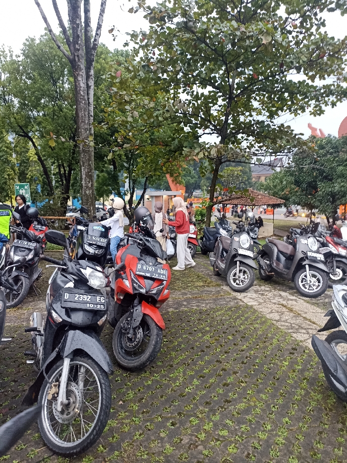 The scene takes place in a parking lot in Jatinangor, Indonesia, located at coordinates -6.931 latitude, 107.776 longitude. There are several parked motorcycles, including a red motorcycle with a license plate that reads "D 6701 ADE" and a black motorcycle with a license plate that reads "D 2392 EG 10-25." Several people are in the scene, including a woman wearing a red hijab and standing by a motorcycle, another woman in a hijab, and a man standing near a motorcycle. The parking lot is covered in a pattern of brick-like pavers. The scene is a casual gathering of people on motorcycles. Several green trees frame the scene. The sky appears cloudy. The environment appears calm and peaceful.